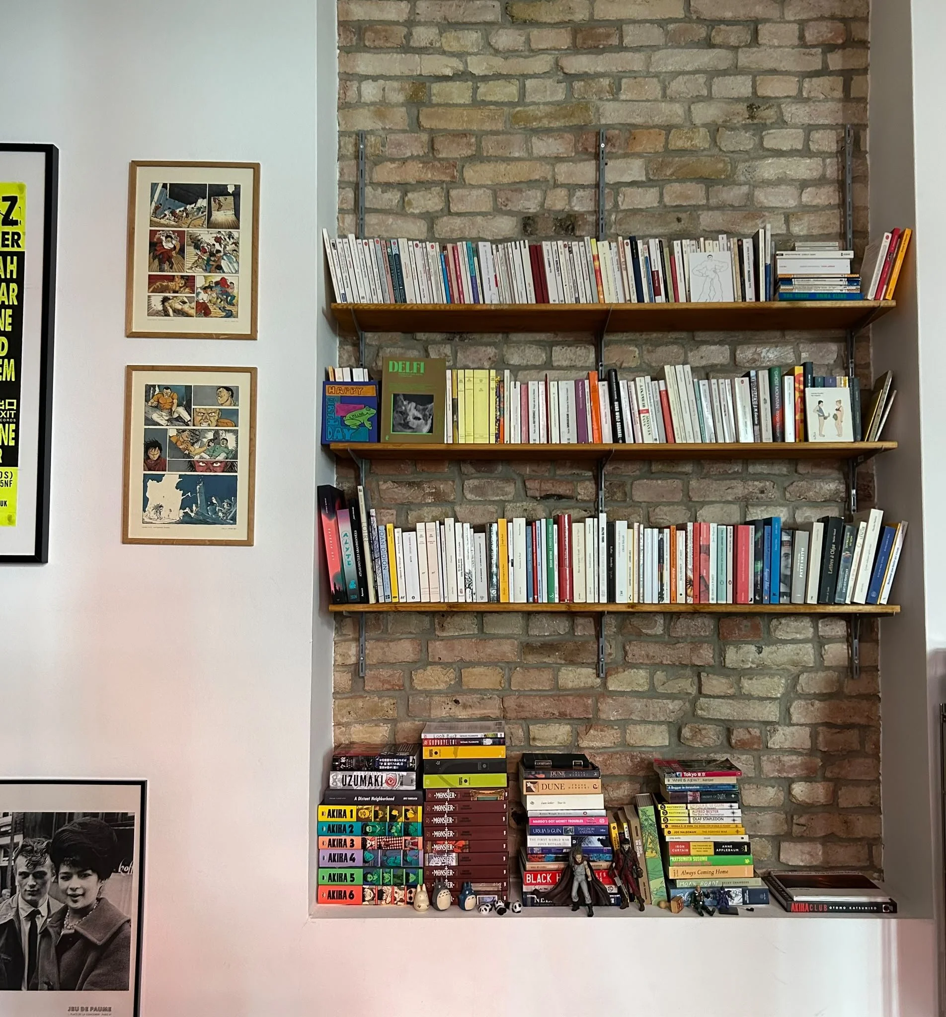 Étagère en bois fixée à un mur en briques, contenant des livres de différentes tailles et couleurs, avec des figurines et des petits objets en dessous. À gauche, une paroi blanche avec des affiches et un poster en noir et blanc.