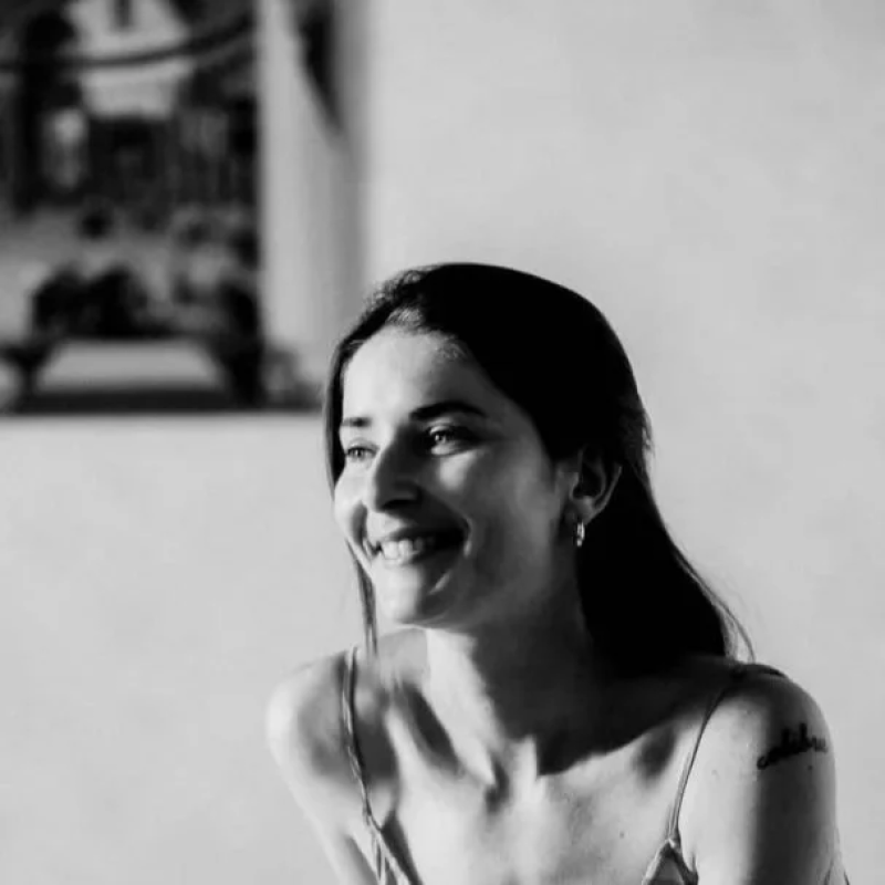 Photo d'Émeline Berton souriante avec des cheveux longs, portant une robe et des boucles d'oreilles, dans un cadre intérieur.