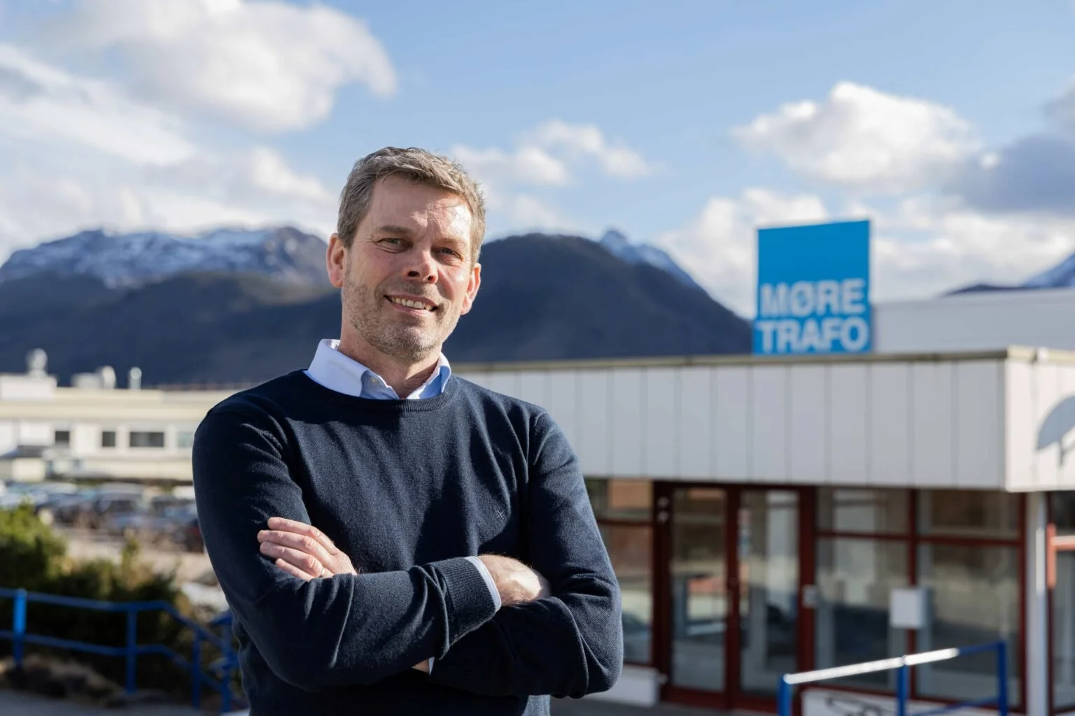Mann med korslagte armer utendørs foran Møre Trafo-bygget med fjell i bakgrunnen.
