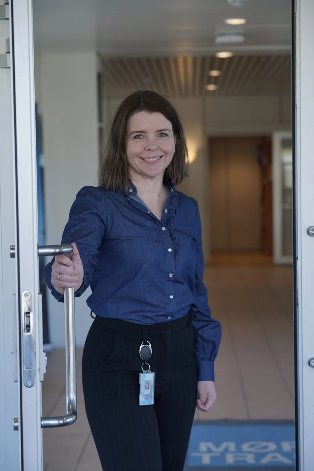 Kvinnelig ansatt i blå skjorte med adgangskort, smilende i døråpning.