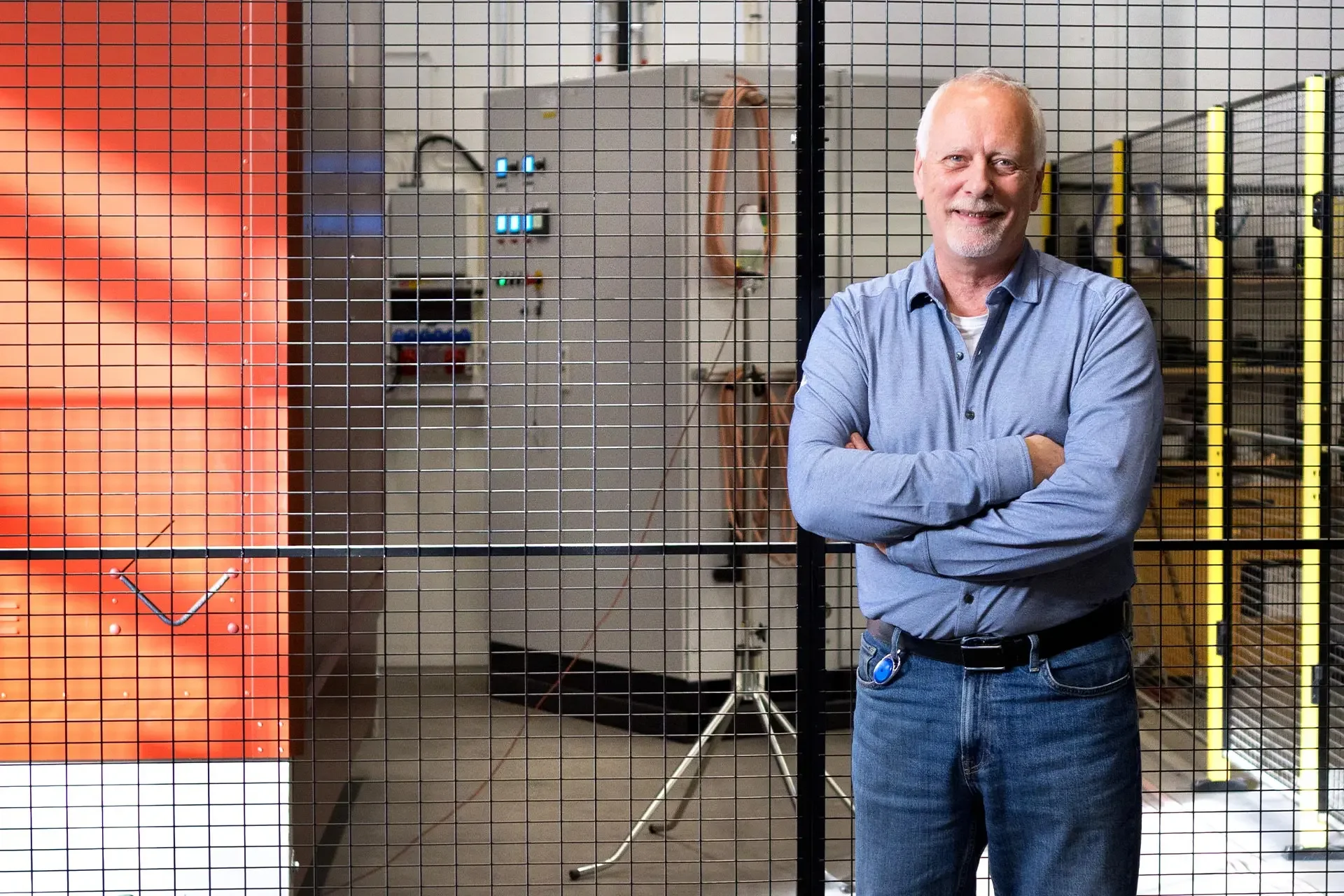 Mann med grått hår og skjegg, korslagte armer, smilende foran teknisk utstyr og oransje panel i industrianlegg.