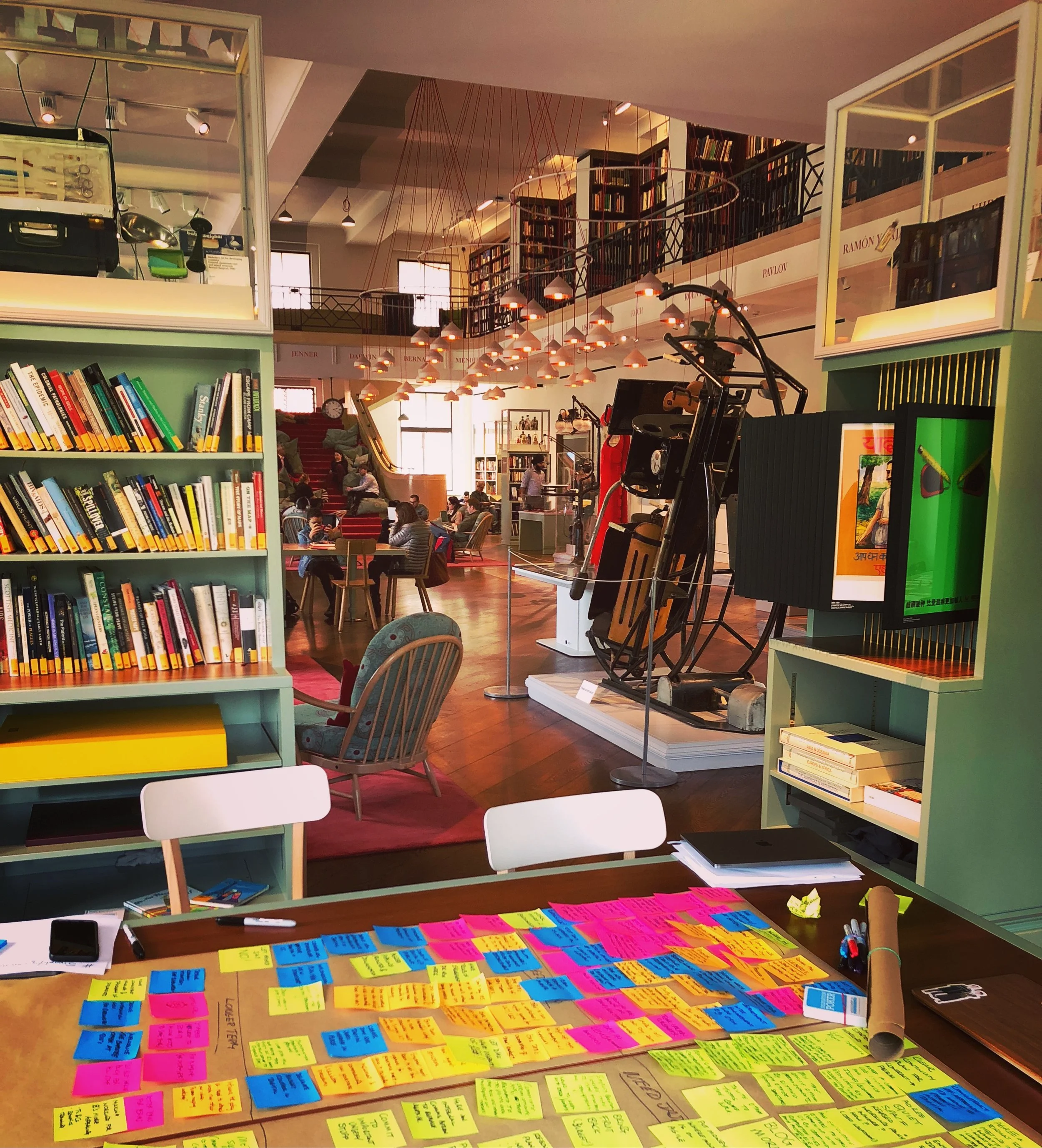 In the Wellcome Collection reading room surrounded books and historical objects and art, a table is covered in neon pink, blue and yellow post it notes