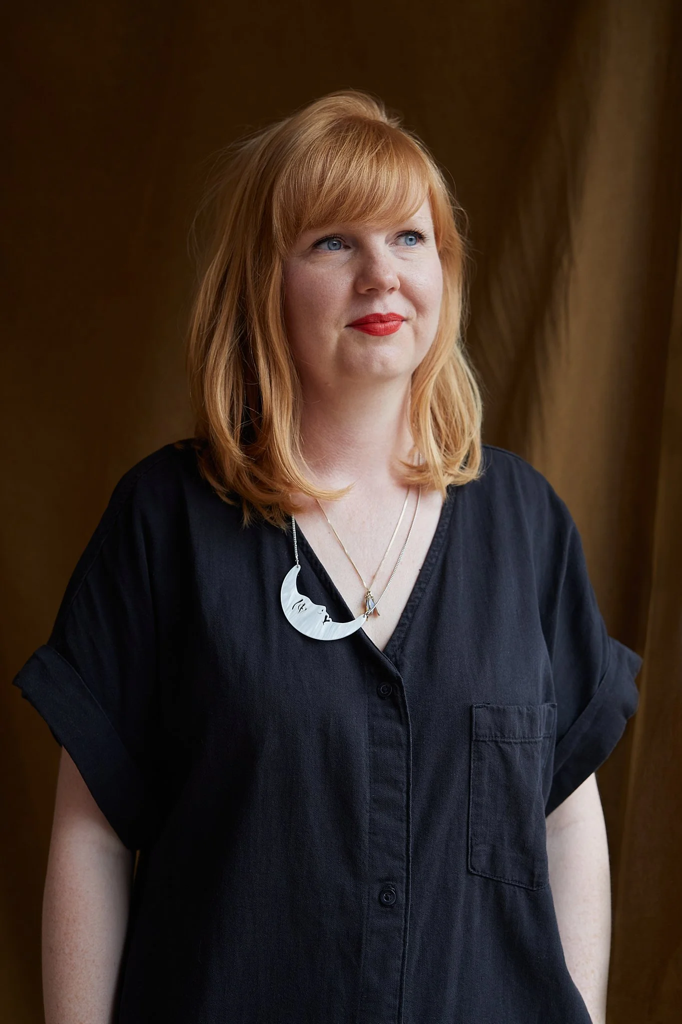 A red haired woman in a black shirt dress with bright red lipstick looks on in the distance, with a moon shaped necklace and rust brown background