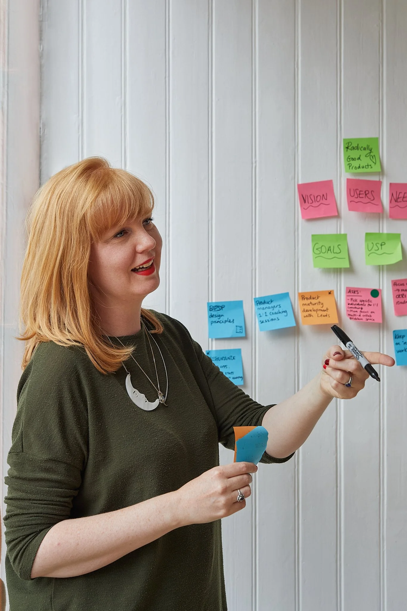 A red haired woman with bright red lipstick and a green top and moon necklace, points at something mid-sentence, holds up a post it that says "Radically good products" while holding a sharpie, smiling behind a wall of neon pink and green post-its