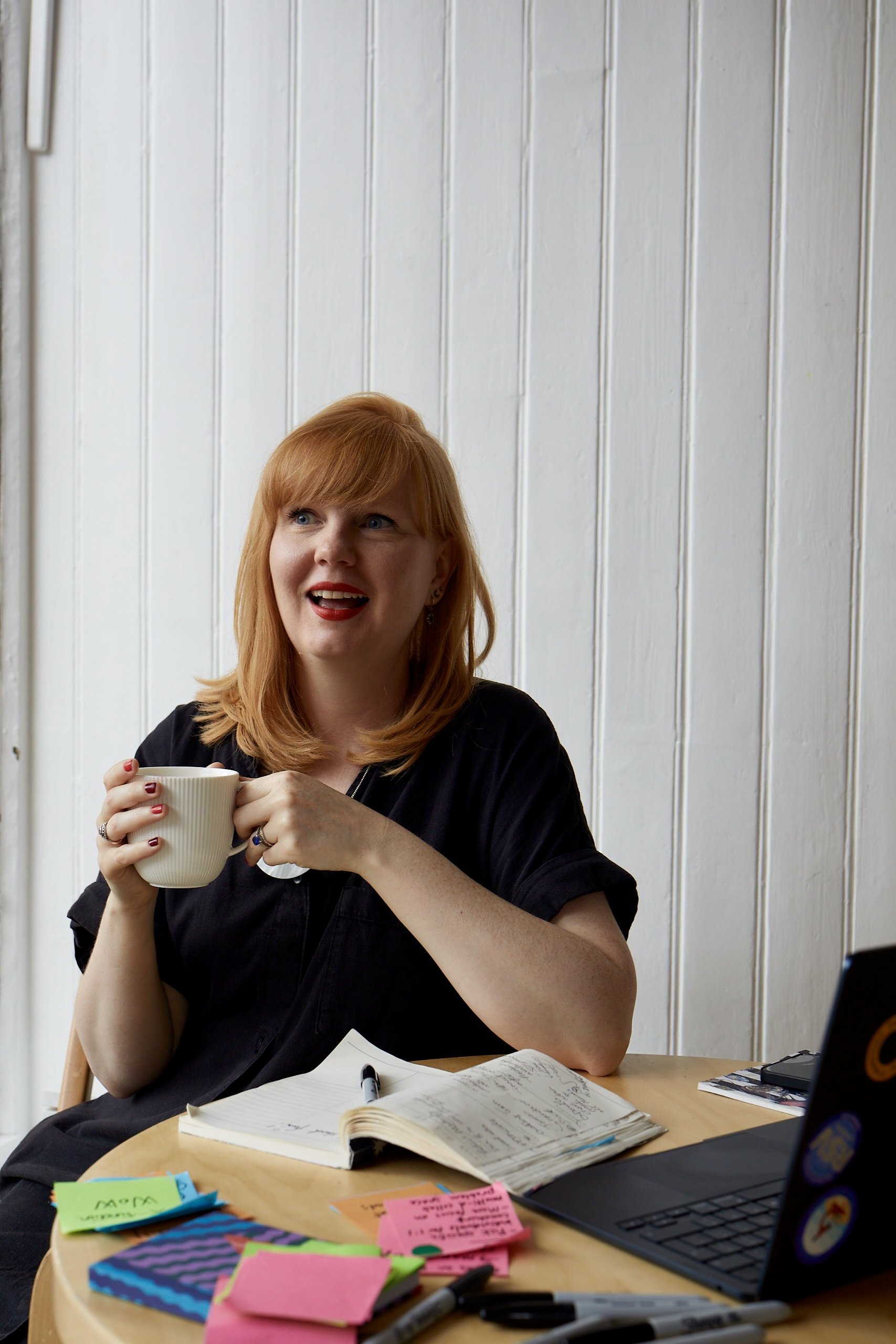 A red haired woman smiles widely with red lipstick, holding a mug of tea and surrounded by post-its a notepad and sharpie and her laptop
