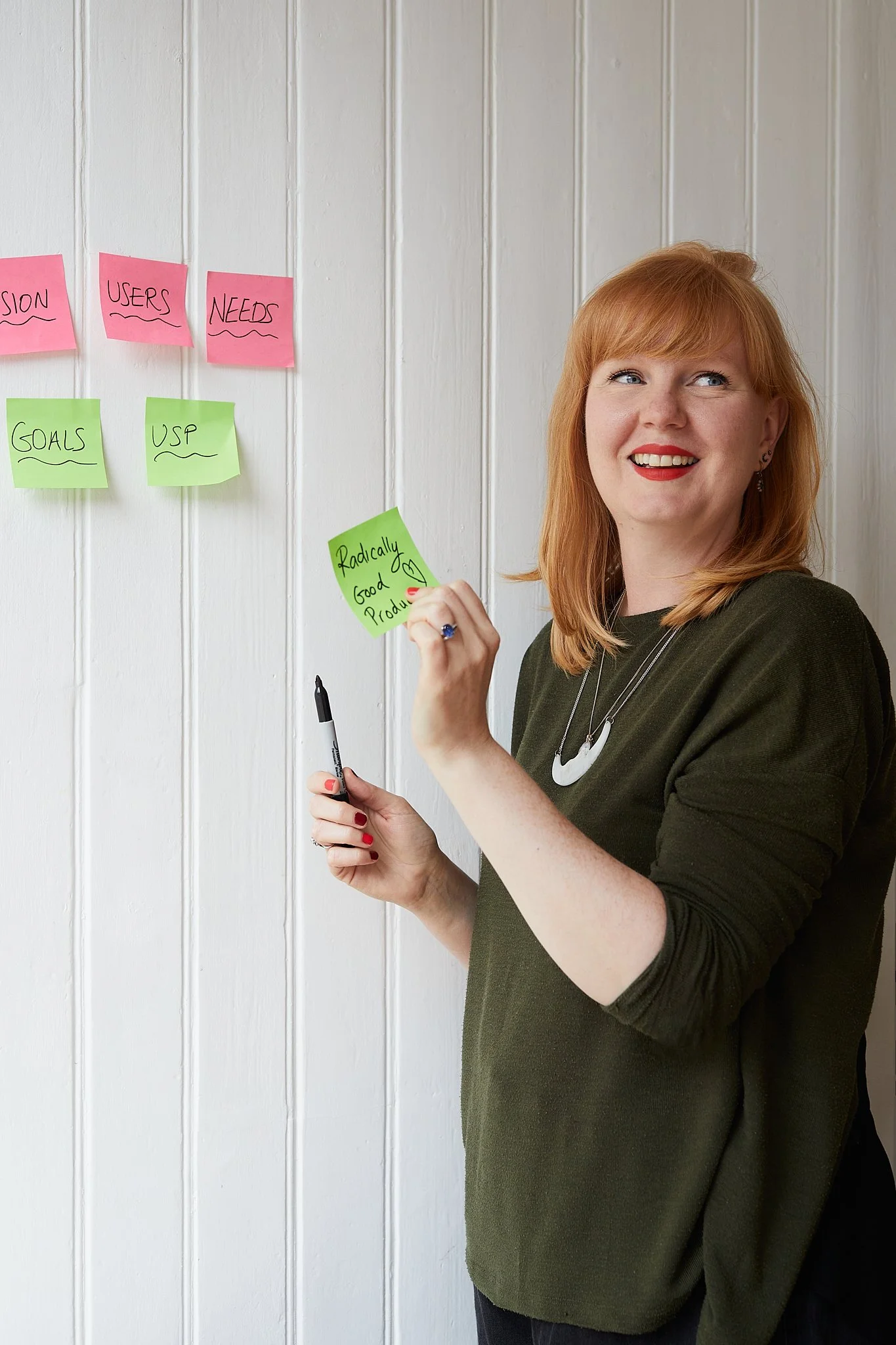 A red haired woman with bright red lipstick and a green top and moon necklace, holds up a post it that says "Radically good products" while holding a sharpie, smiling behind a wall of neon pink and green post-its