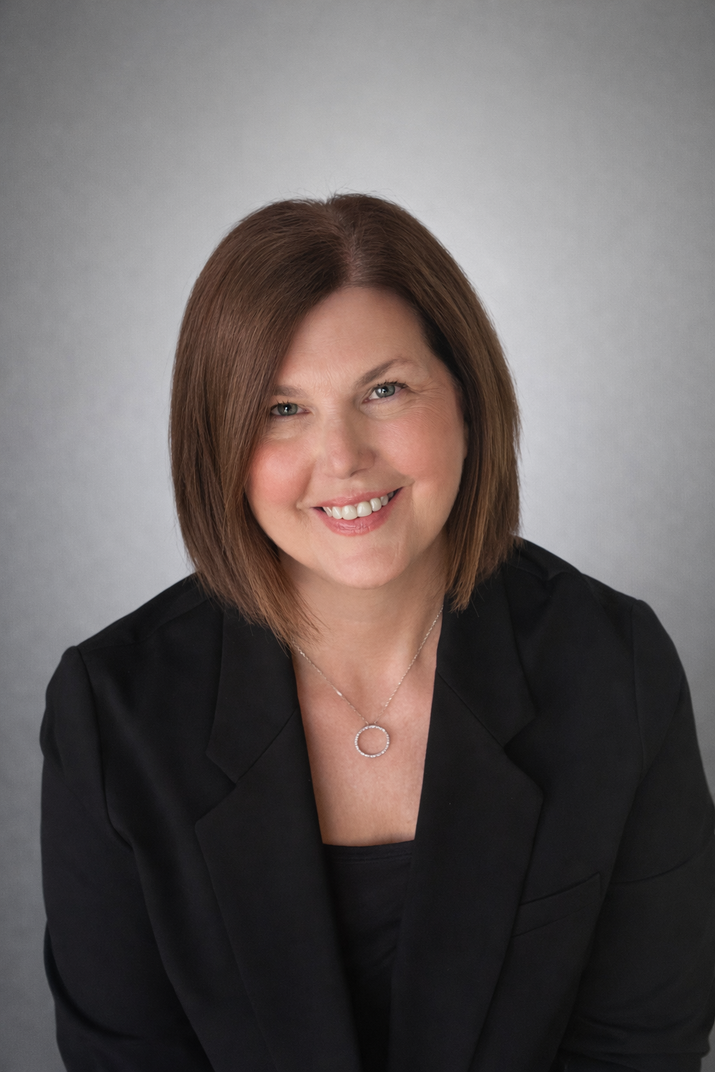 A woman with shoulder-length brown hair and blue eyes smiling, wearing a black blazer and a silver necklace, against a gray background.
