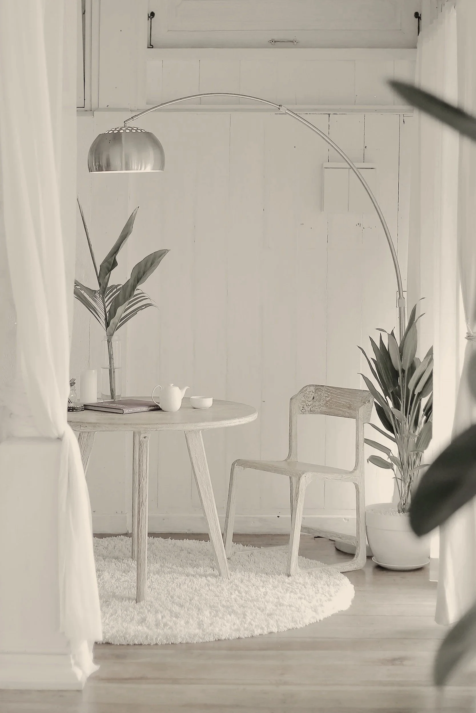 A minimalist, light-colored room with a round wooden table, a wooden chair, large potted plants, a white rug, and a silver arc floor lamp.