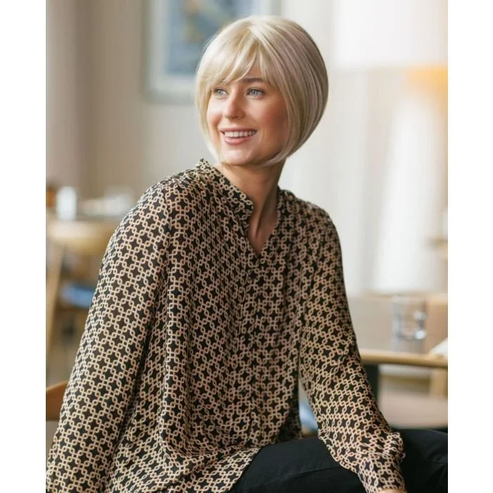 Woman with short blonde hair smiling, wearing a patterned blouse, seated indoors with a blurred background.