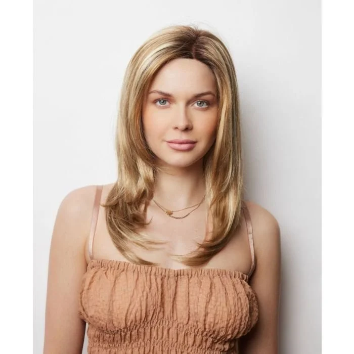 A young woman with long blonde hair, blue eyes, and fair skin standing against a white wall, wearing a peach-colored top and a gold necklace.