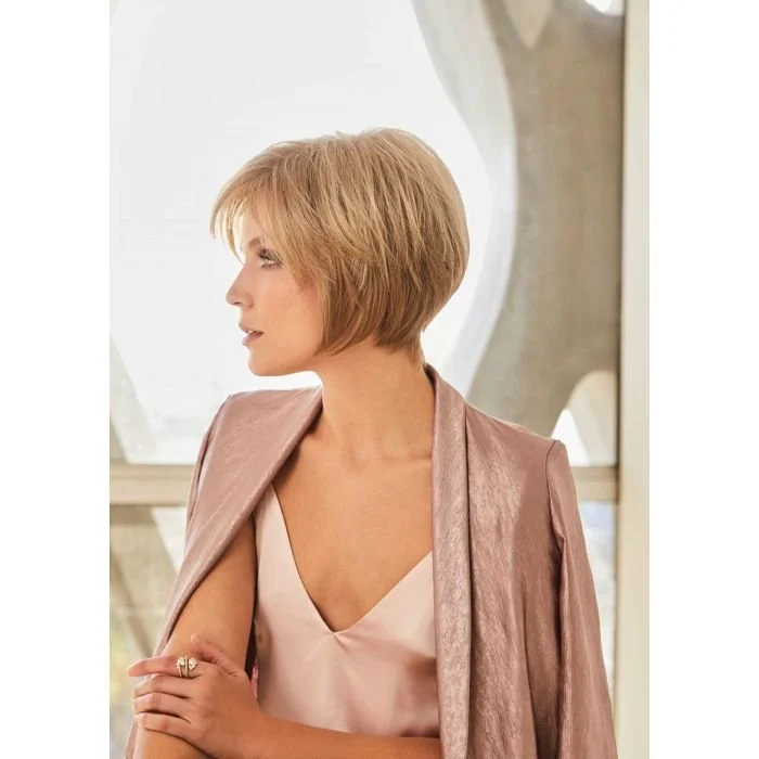 A woman with short blonde hair looking to the side, wearing a pink satin dress and a matching shawl, standing in front of a large window.