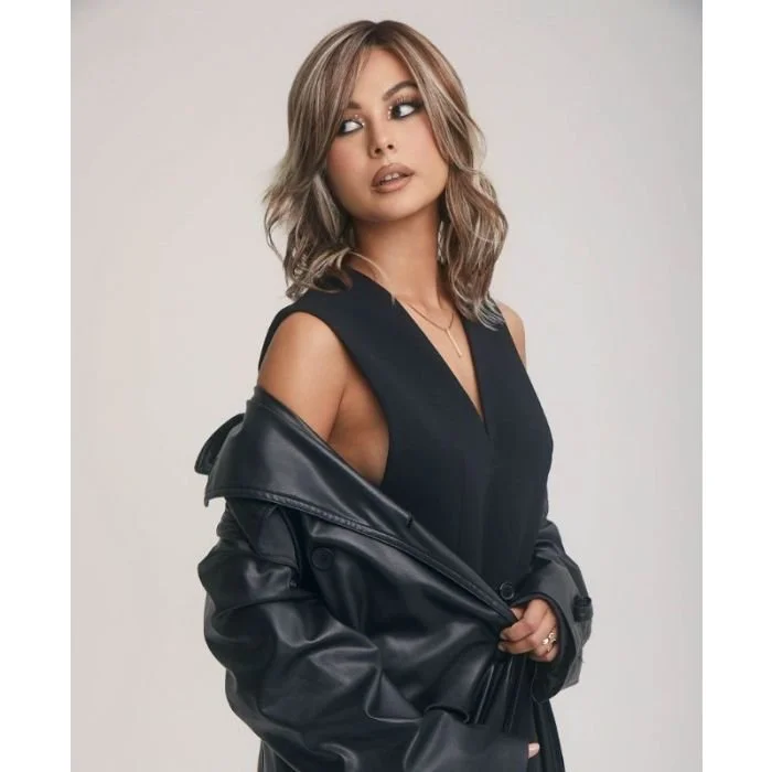 A young woman with shoulder-length wavy hair and light makeup, looking to her right, wearing a black sleeveless top and a black leather jacket draped over her shoulders, standing against a plain light gray background.