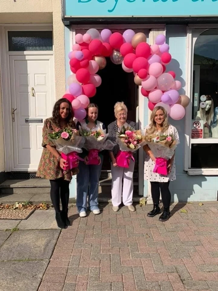 Four women standing outside a shop, holding bouquet of flowers, in front of pink and purple balloon arch for celebration.