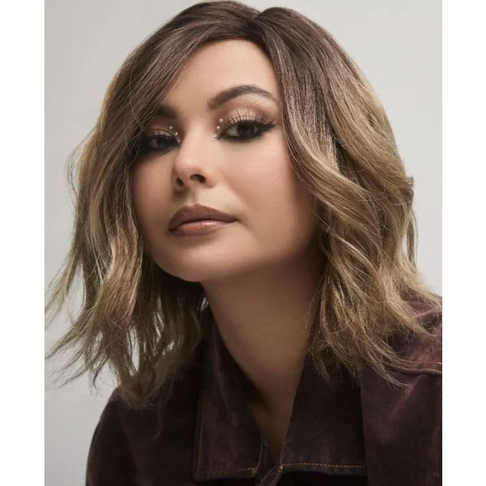 Close-up of a young woman with shoulder-length wavy brown hair and makeup featuring prominent eye details and neutral lipstick, wearing a dark jacket against a plain background.