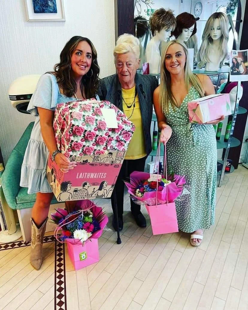 Three women standing inside a hair salon holding gifts and flowers, with mannequin heads with wigs in the background.
