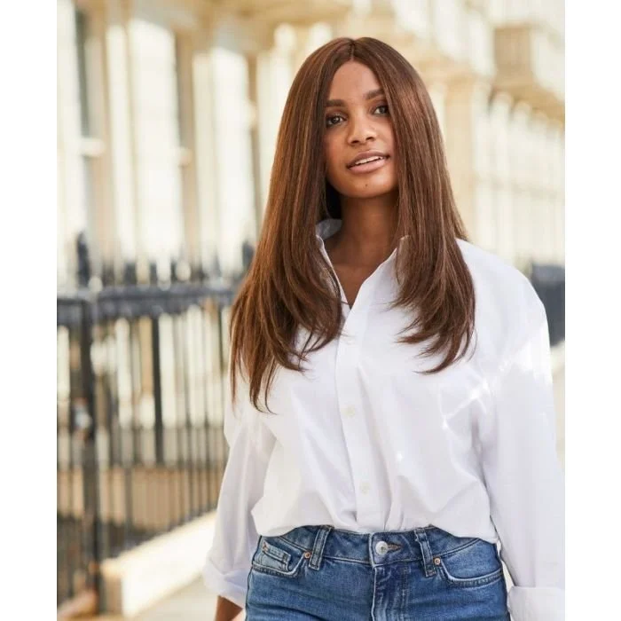 Young woman with long brown hair wearing a white shirt and jeans standing outdoors near a railing with a blurred background of porch columns.