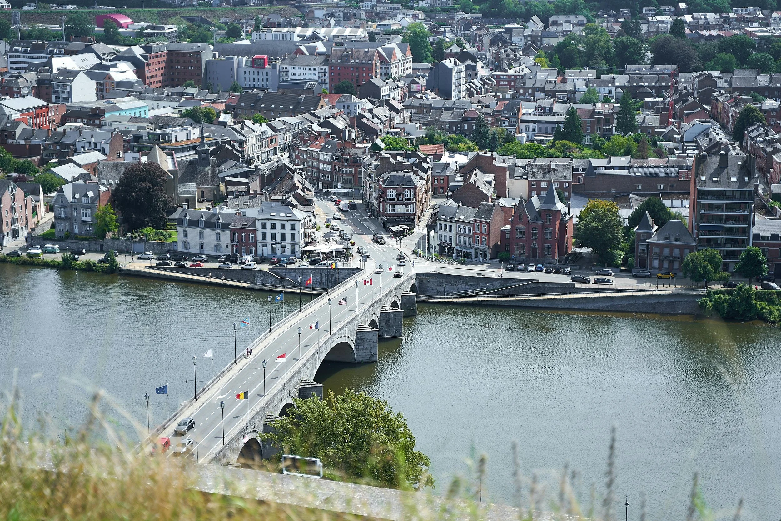 Vue aérienne d'une ville avec un fleuve, un pont en pierre avec des drapeaux, et des bâtiments résidentiels et commerciaux.