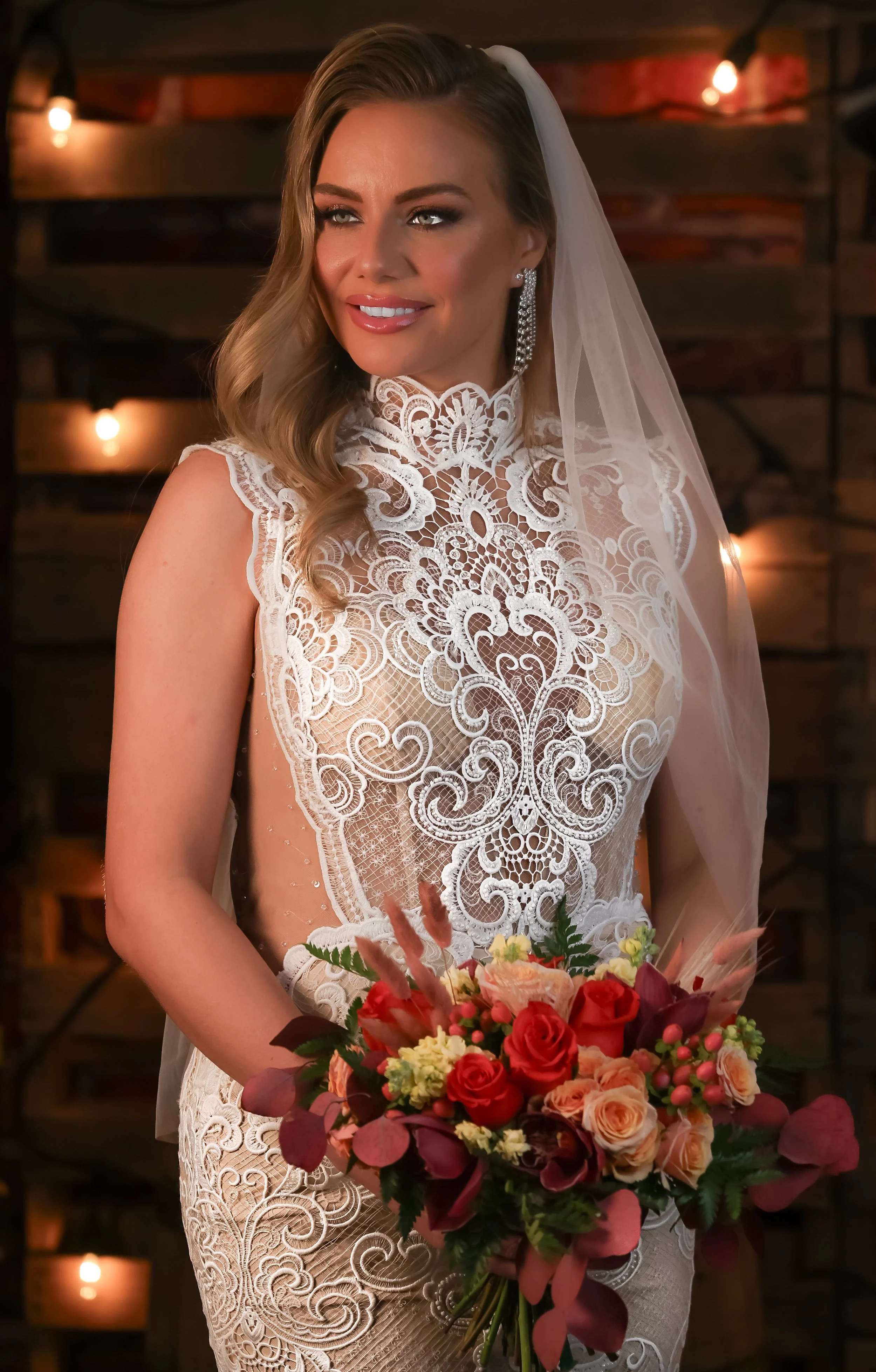 A bride wearing a detailed white lace wedding dress holding a vibrant bouquet of flowers, with a wooden backdrop and warm string lights.