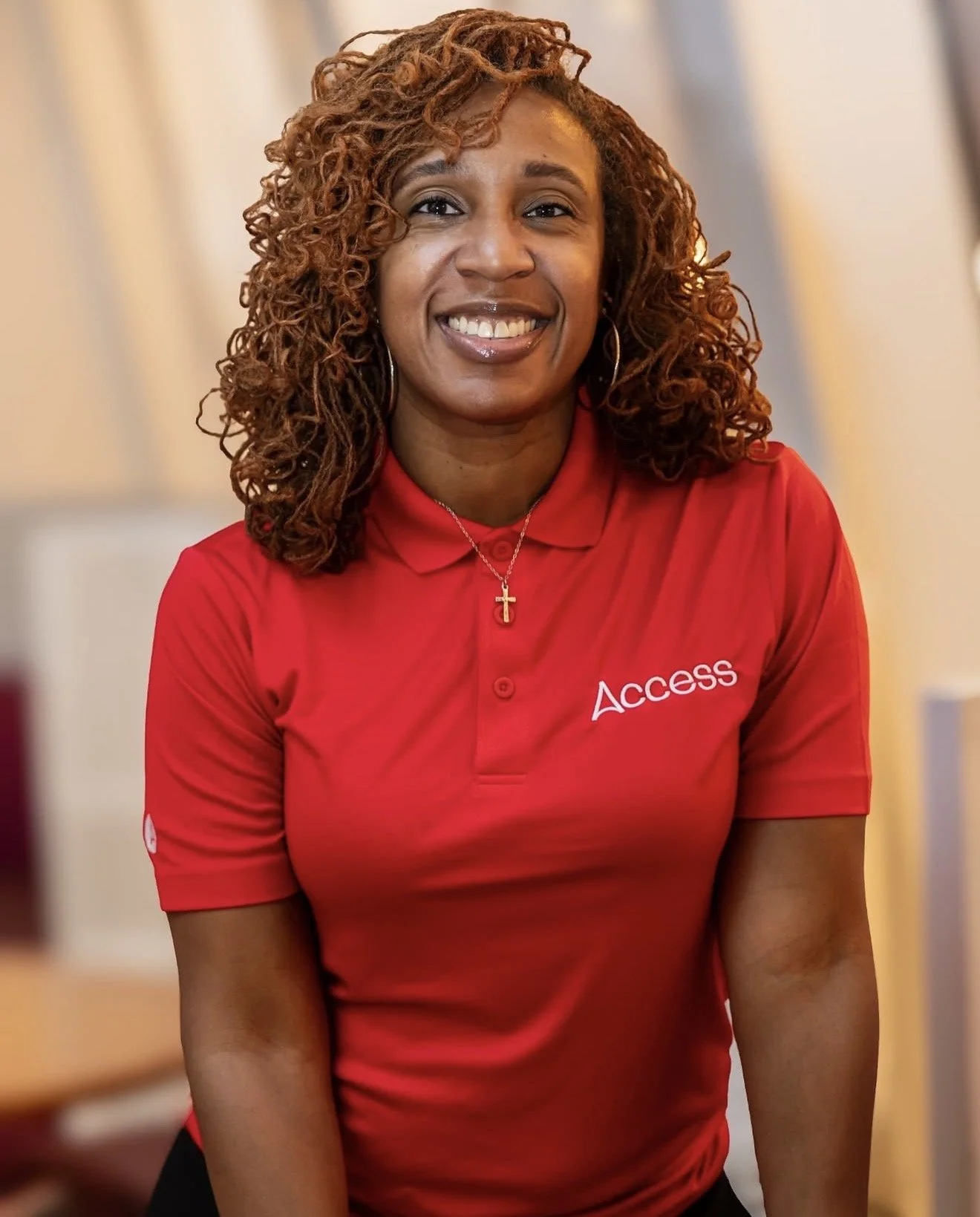 A woman with curly hair wearing a red polo shirt with the word 'Access' and a cross necklace, smiling in an indoor setting.