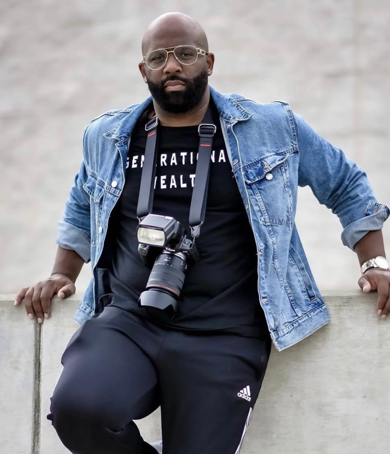 Man with glasses and a beard wearing a denim jacket and black Adidas sweatpants, sitting on a concrete ledge with camera around neck.