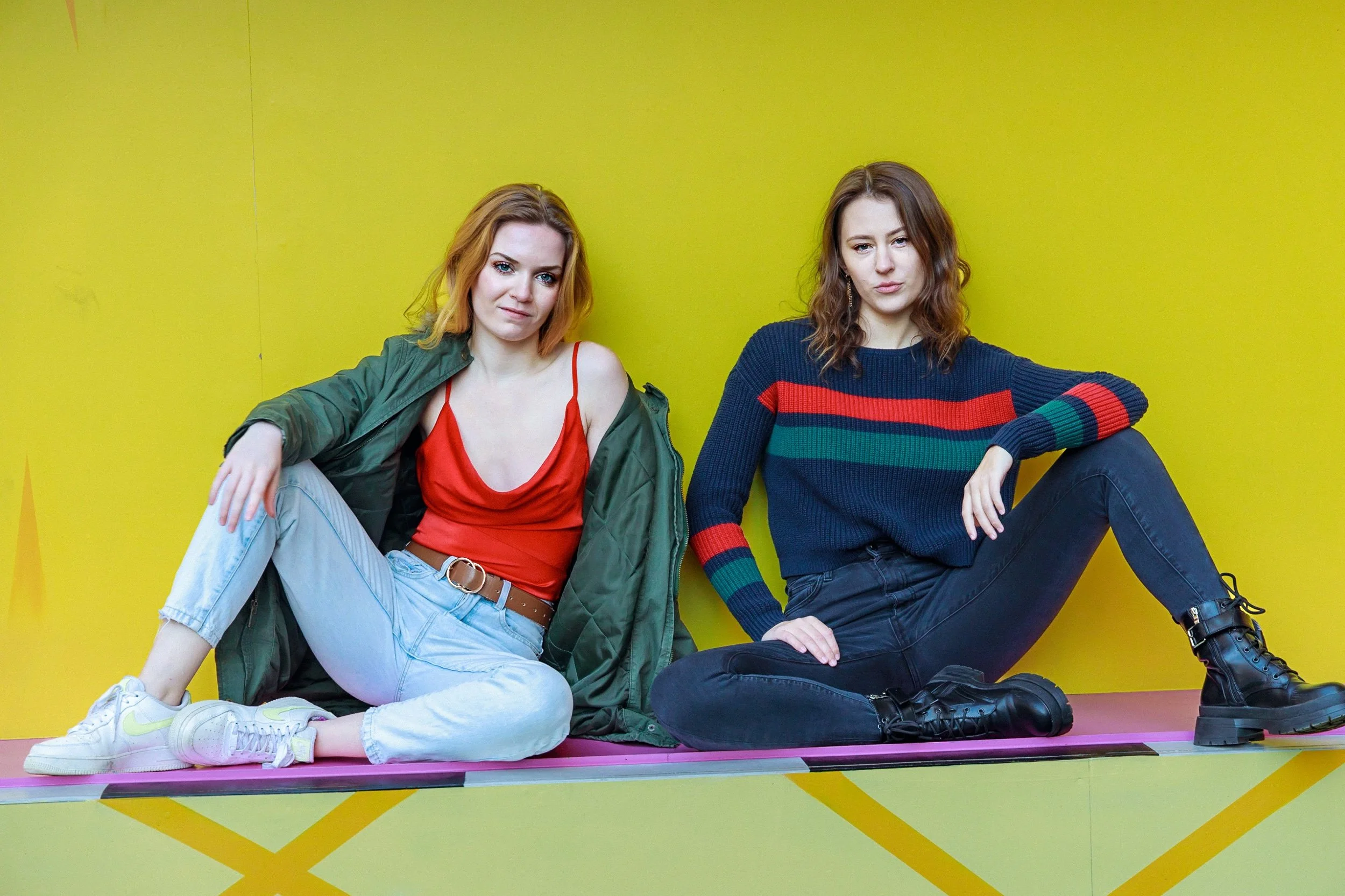 Two young women sitting on a pink surface against a yellow background. One has red hair, wearing a red camisole, green jacket, and light jeans. The other has brown hair, wearing a dark sweater with red and green stripes and black pants.