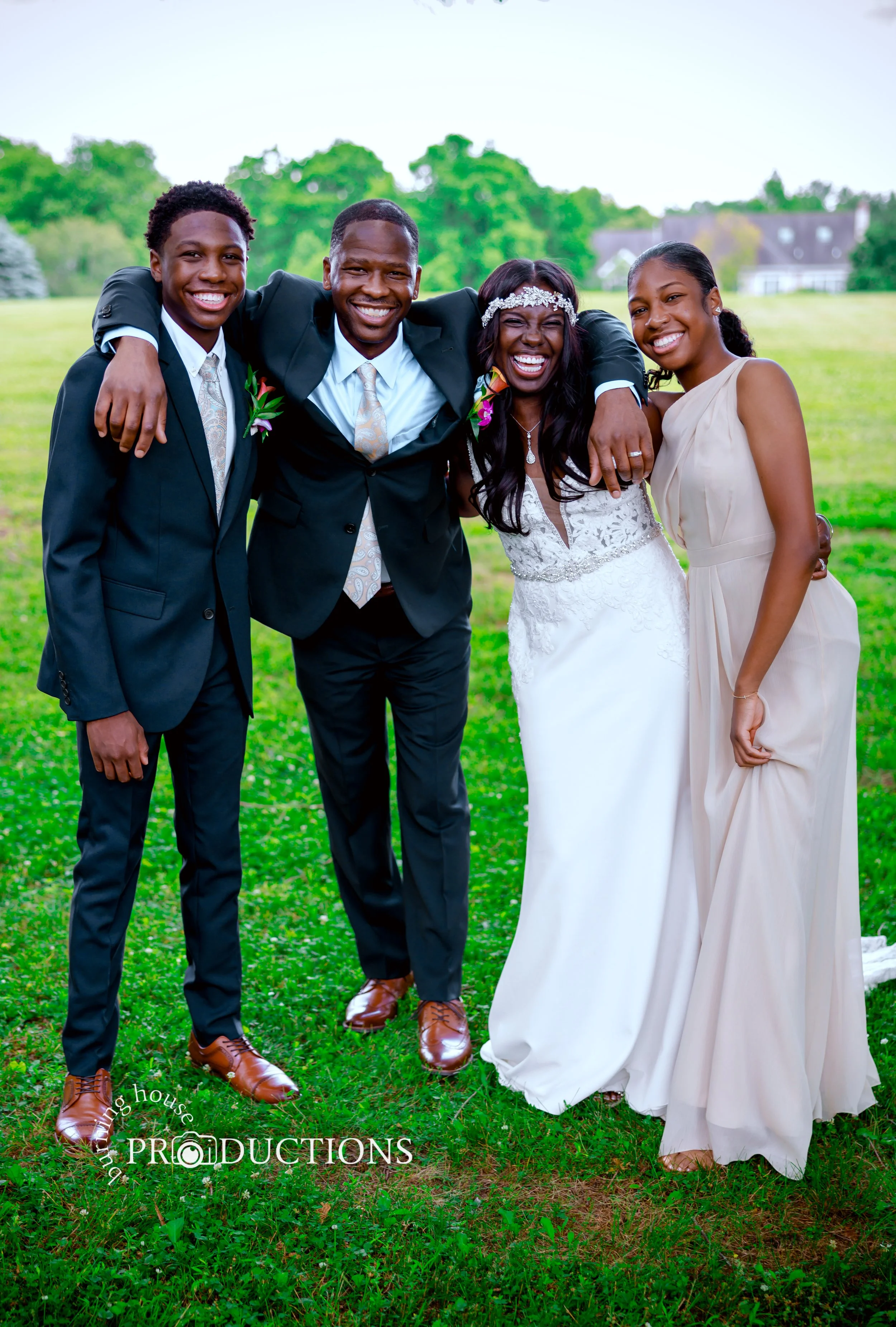 A happy group of five people, including a bride and groom, smiling and hugging outdoors in a grassy area with trees and homes in the background.