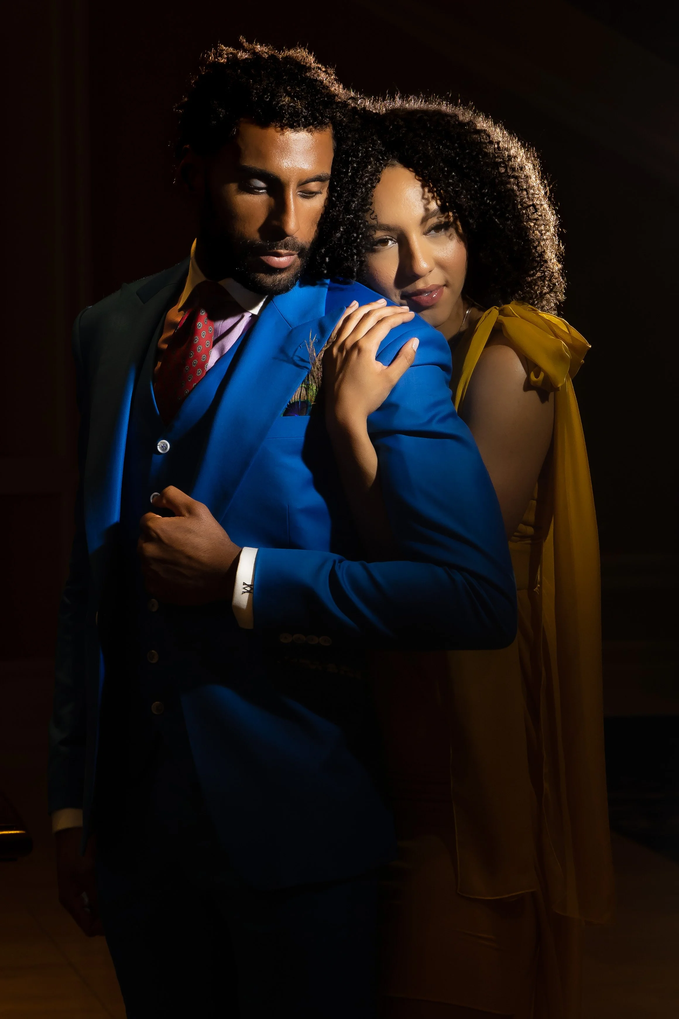 A man and woman in stylish clothing pose closely together in a dark setting, with dramatic lighting highlighting their faces and outfits.