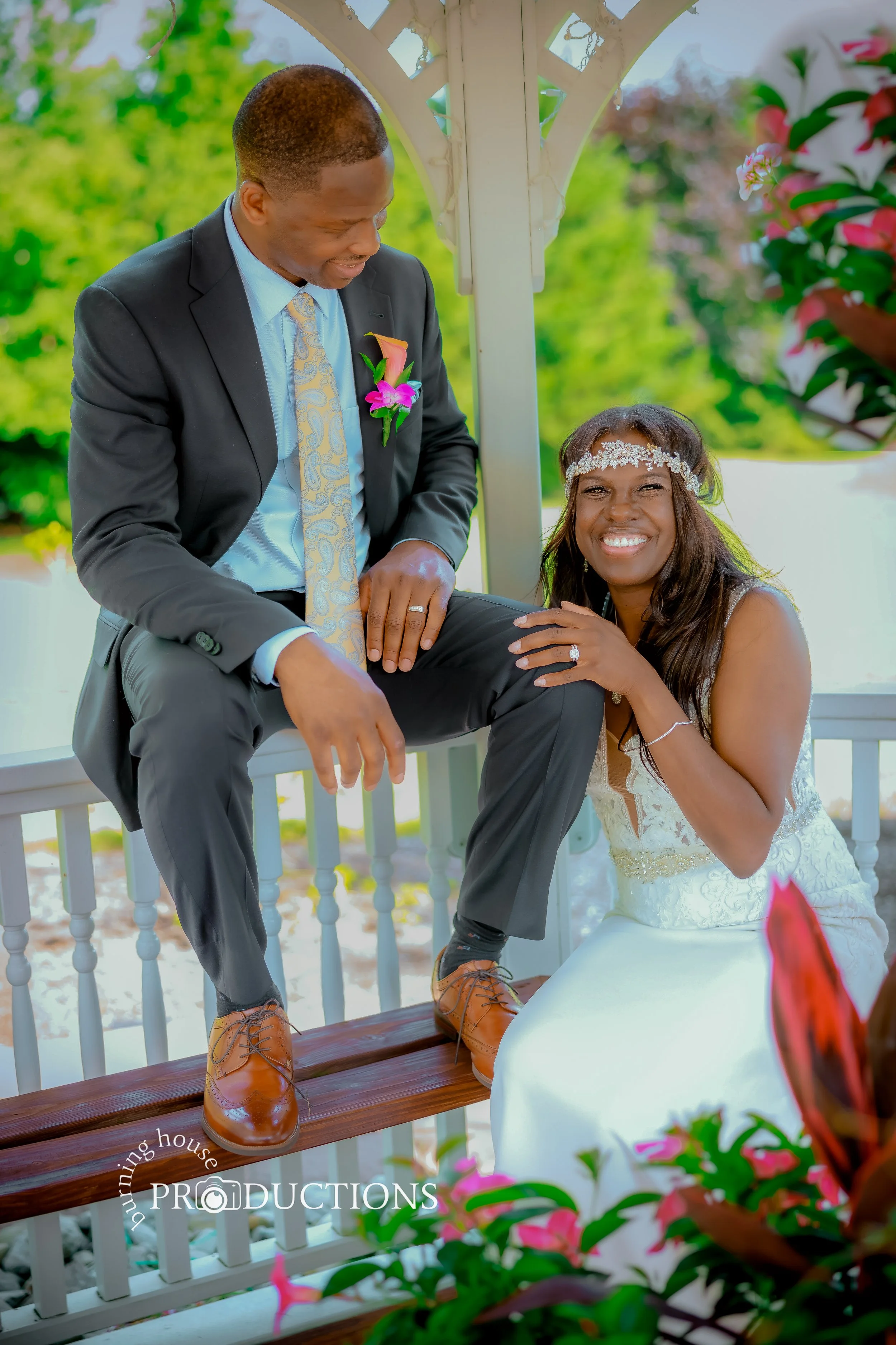 A smiling woman in a white wedding dress and headband poses with a man in a black suit, light blue shirt, and patterned tie. The man has one foot on a bench and the woman kneels beside him, both showing engagement rings. They are in a gazebo with pin