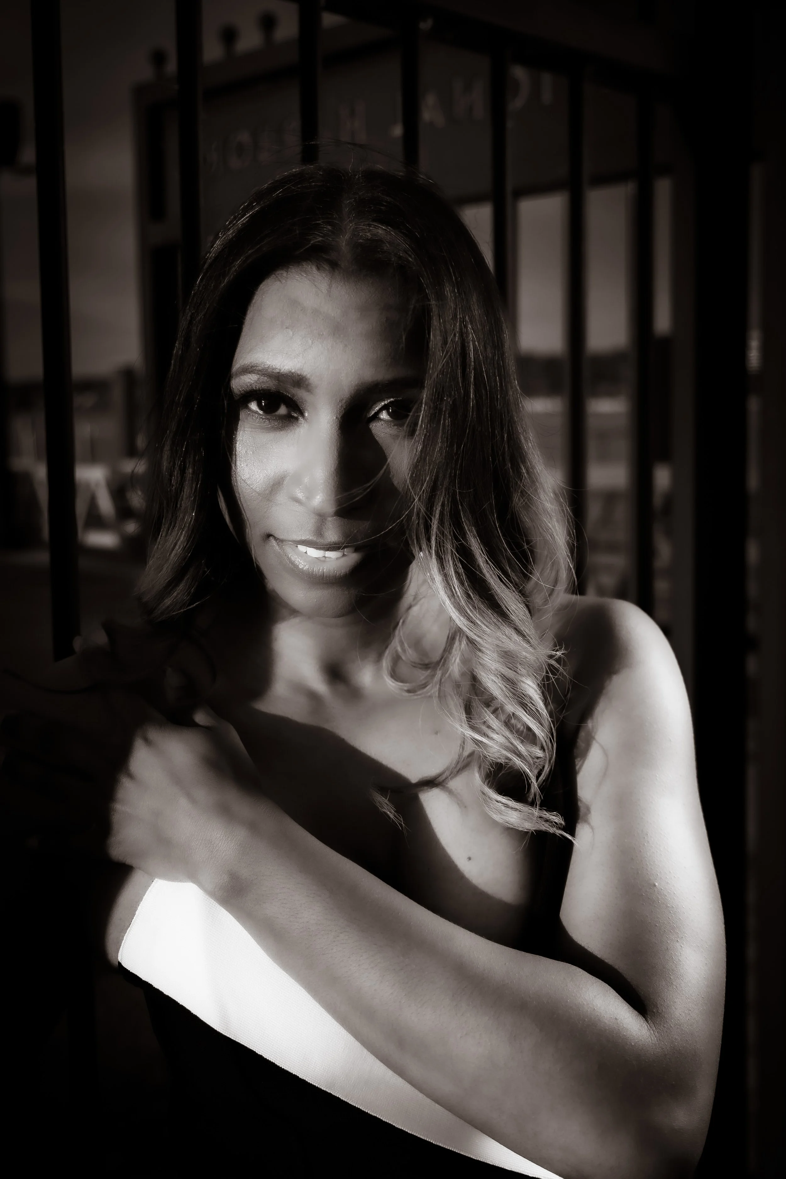 A woman with shoulder-length wavy hair, partially show her face and shoulder, arm crossing her body, in a dimly lit setting created by sunlight, with a grid of metal bars in the background.