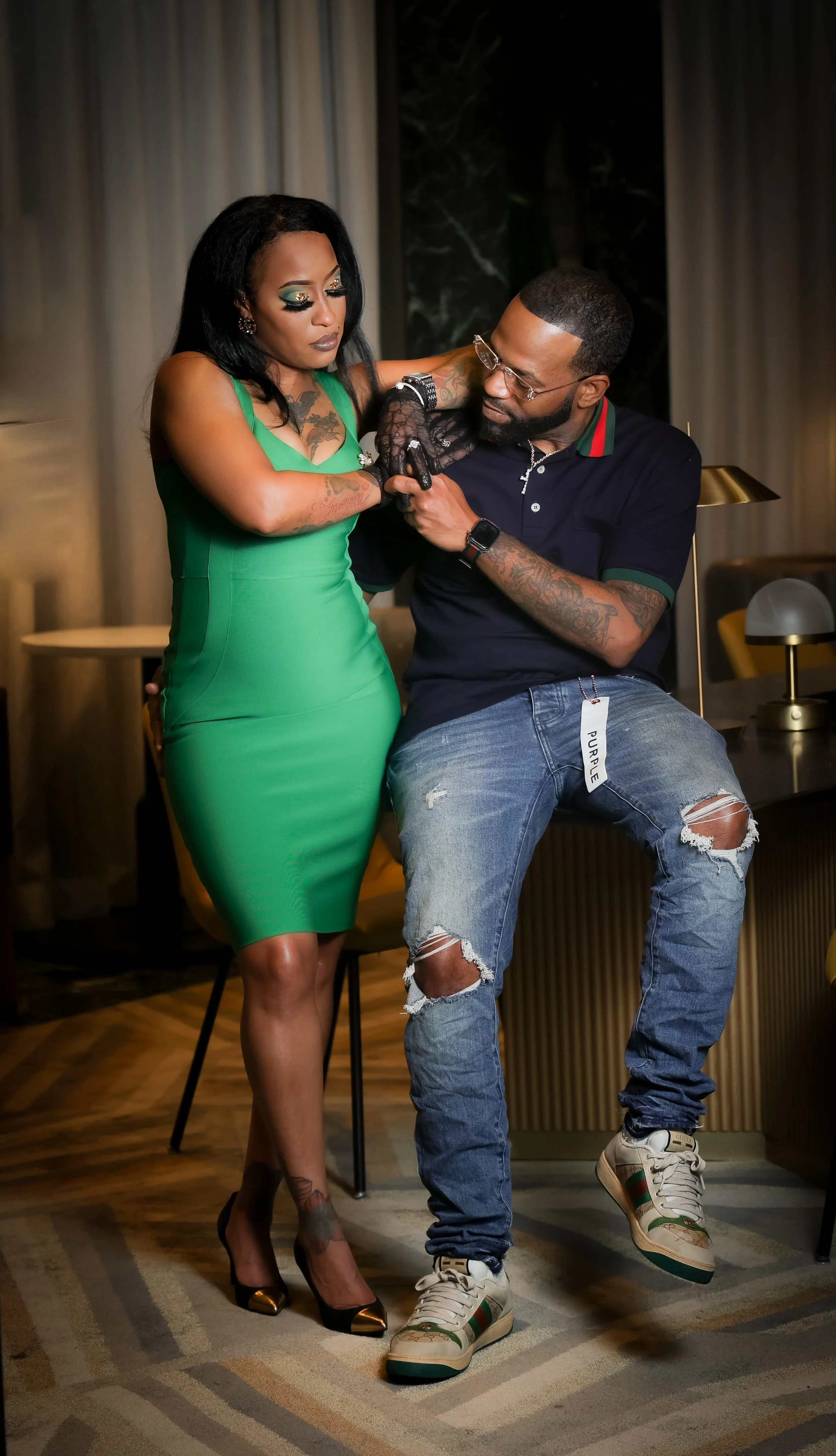 A woman in a green dress and high heels stands next to a man with tattoos, glasses, and a beard, wearing ripped jeans and sneakers, in an indoor setting. The man is sitting on a table while the woman is engaging with him.