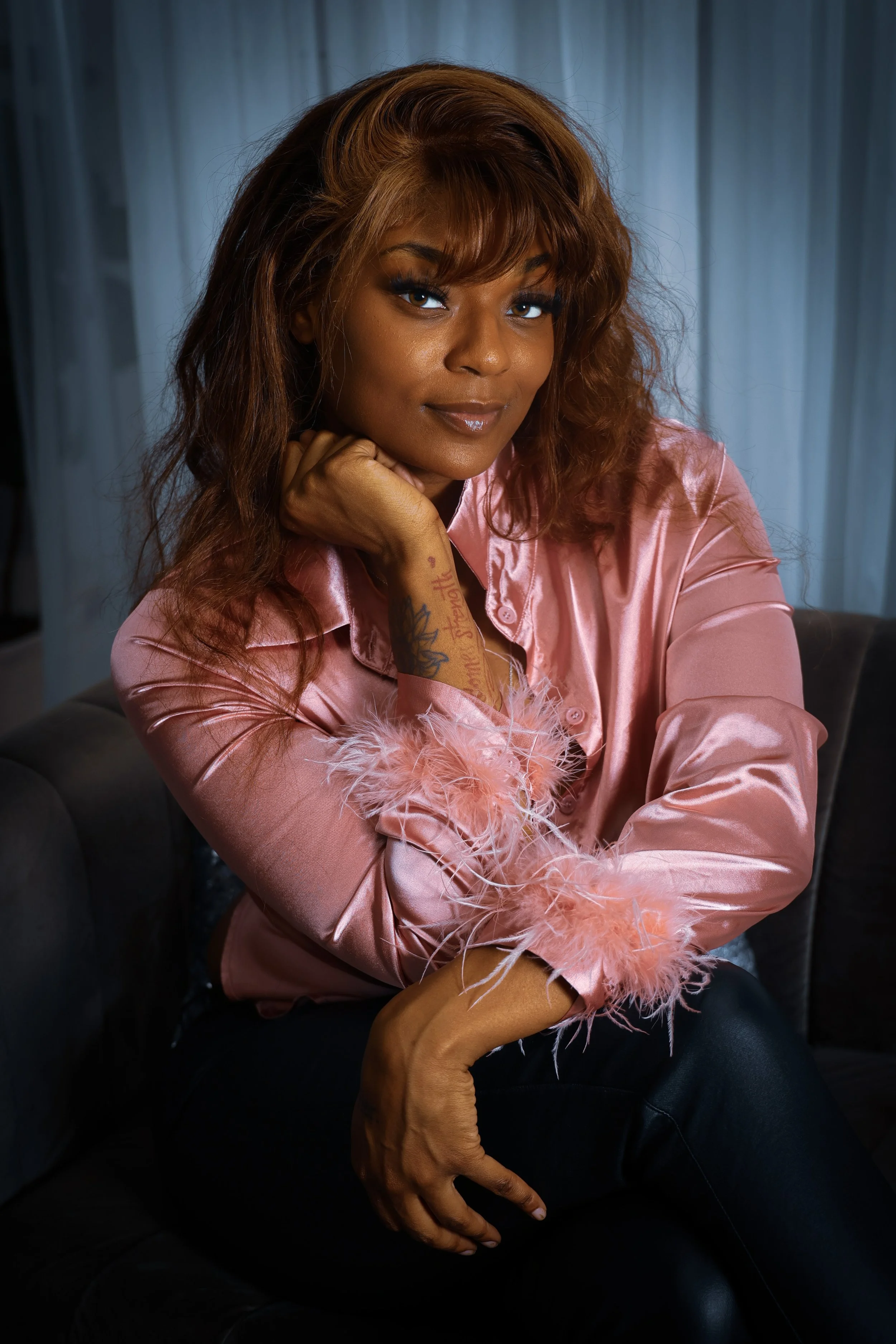 A woman with shoulder-length wavy brown hair and a tan skin tone, wearing a pink satin shirt with feathered cuffs, sitting on a dark-colored sofa in front of a light-colored curtain backdrop.