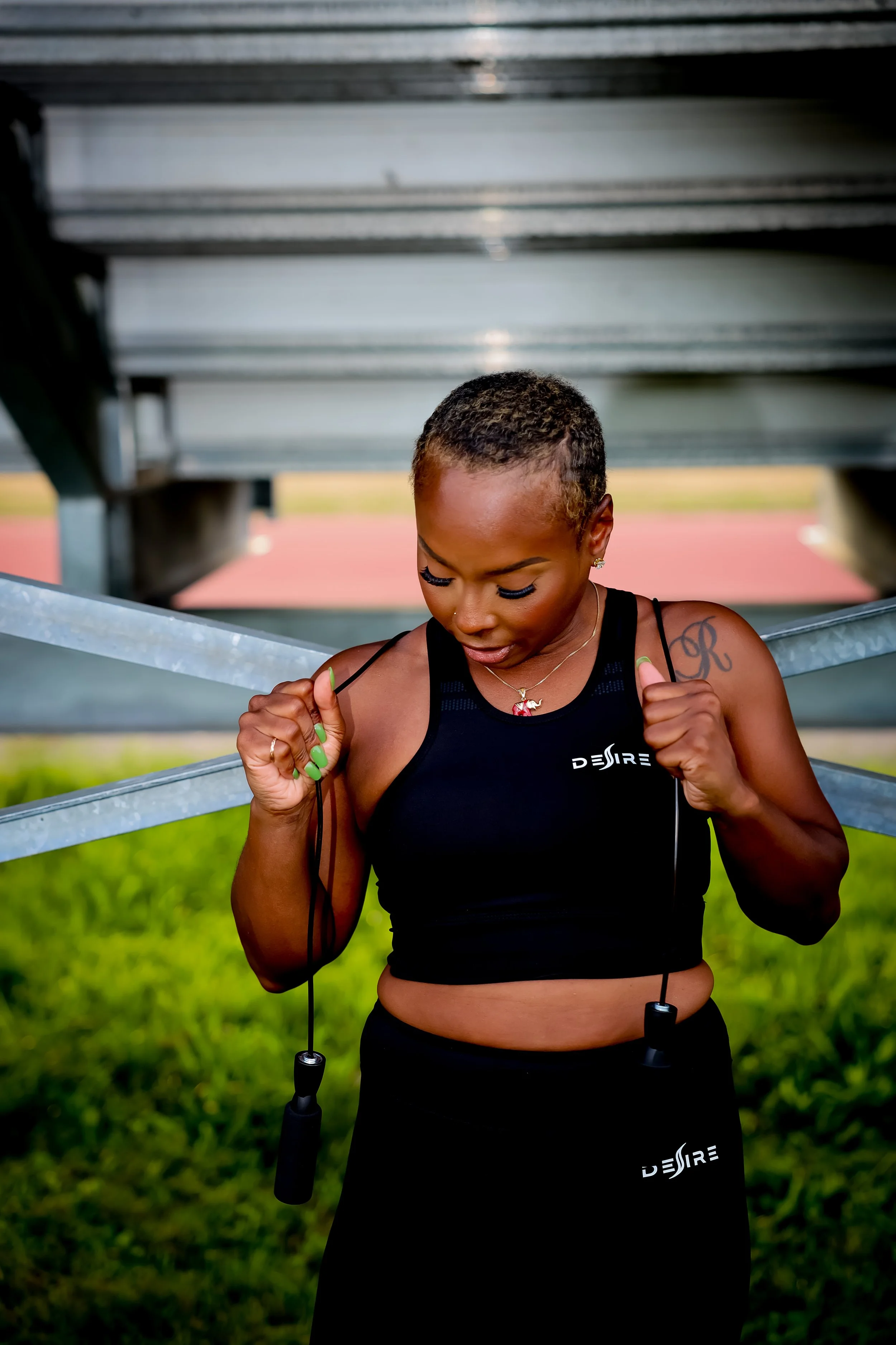 A woman with short hair, wearing a black sports top and skirt, holding a jump rope, standing outdoors on grass under bleachers.