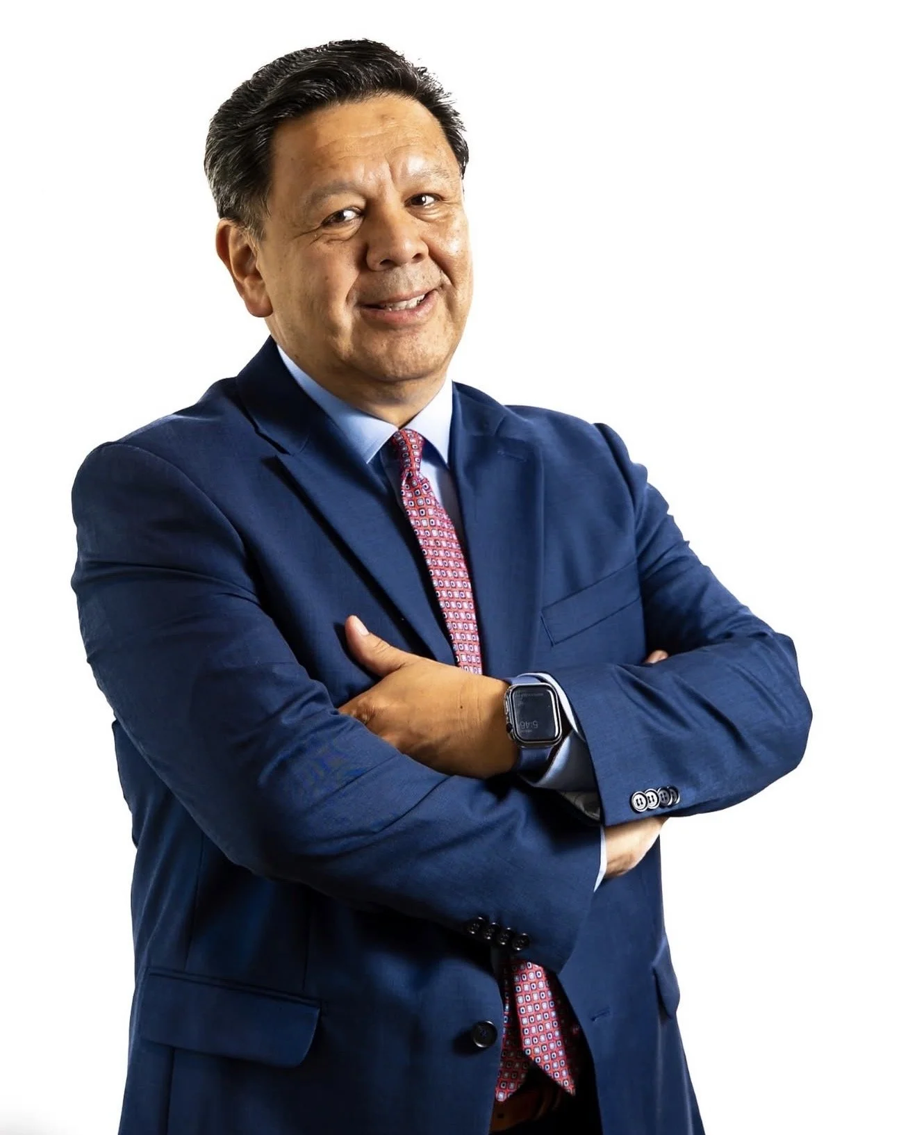 A man in a blue suit with a patterned red tie, crossing his arms and smiling, wearing a smartwatch on his left wrist, against a white background.