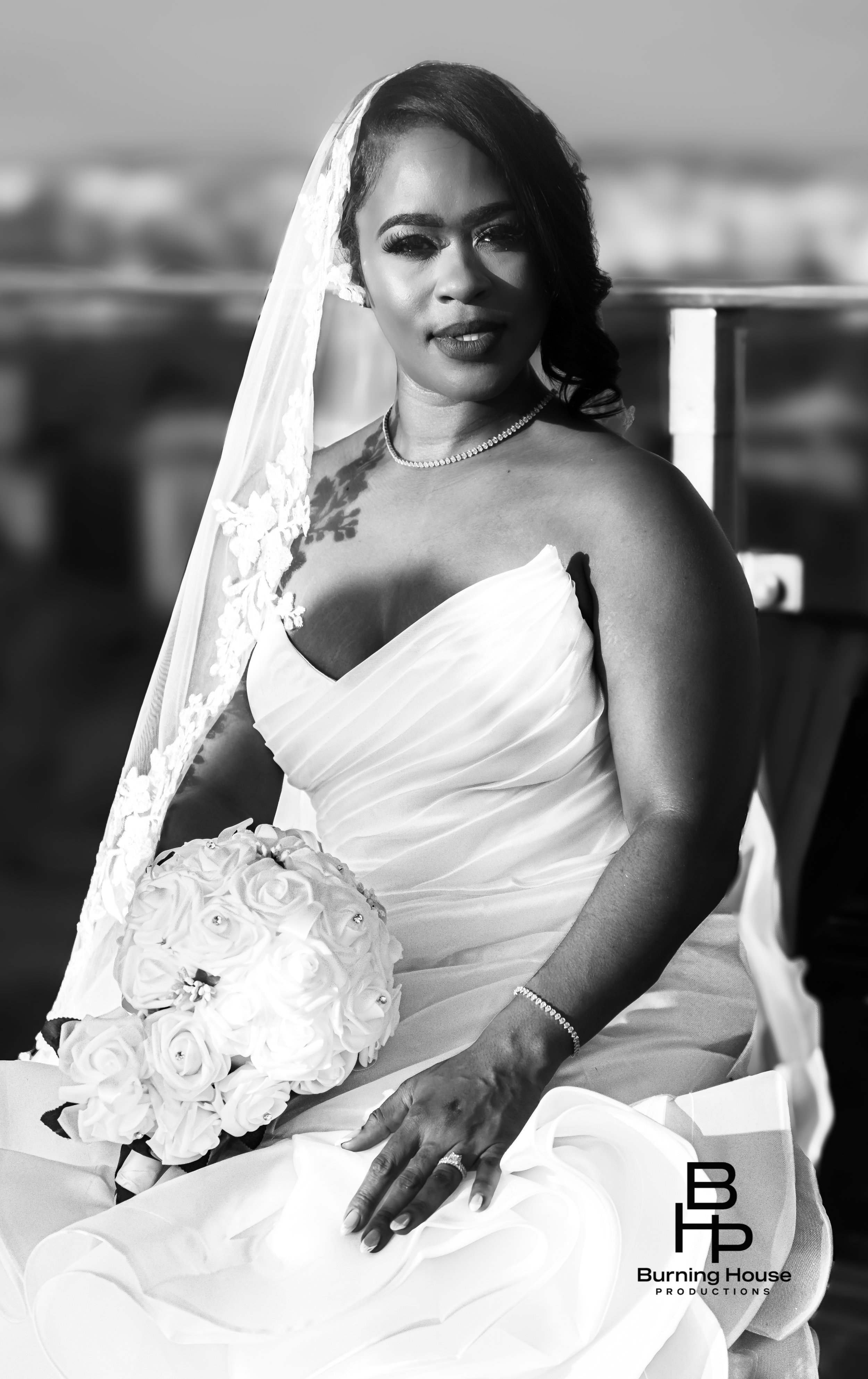 Black-and-white photo of a woman in a wedding dress, holding a bouquet of roses, sitting outdoors, wearing a veil, necklace, bracelet, and ring, with a cityscape blurred in the background.