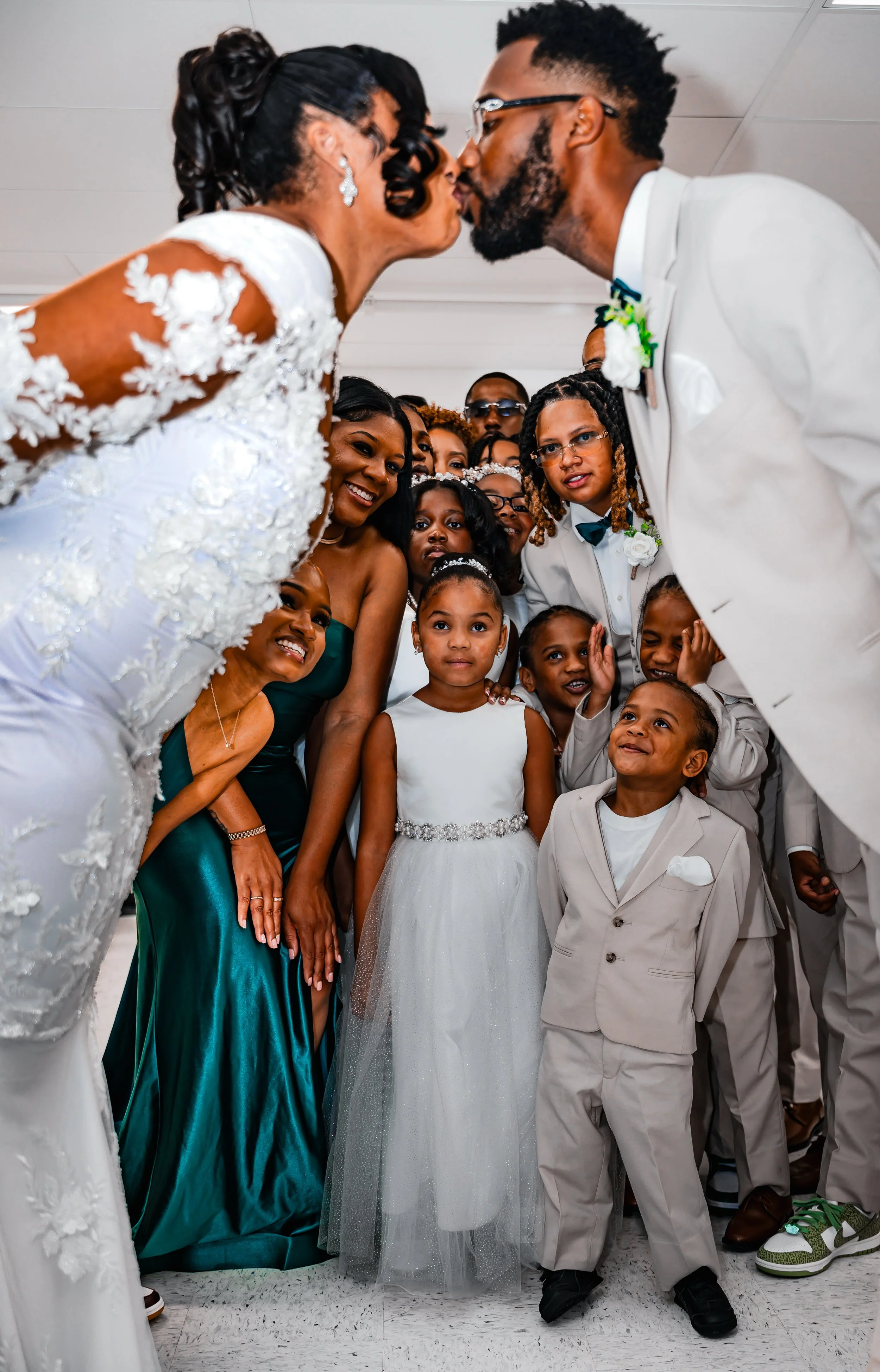 A bride and groom are leaning in to kiss each other at their wedding reception, surrounded by a group of children and adults watching and smiling.