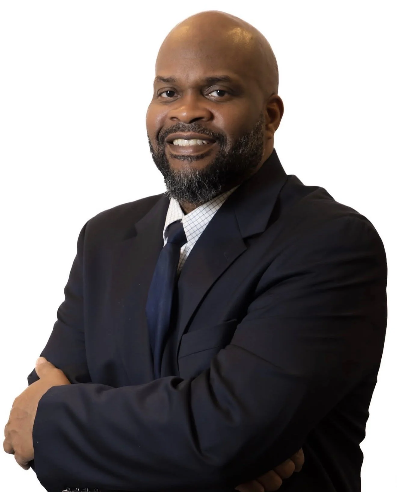 A bald African American man with a beard, wearing a dark suit, white shirt, and dark tie, standing with arms crossed against a white background and smiling.