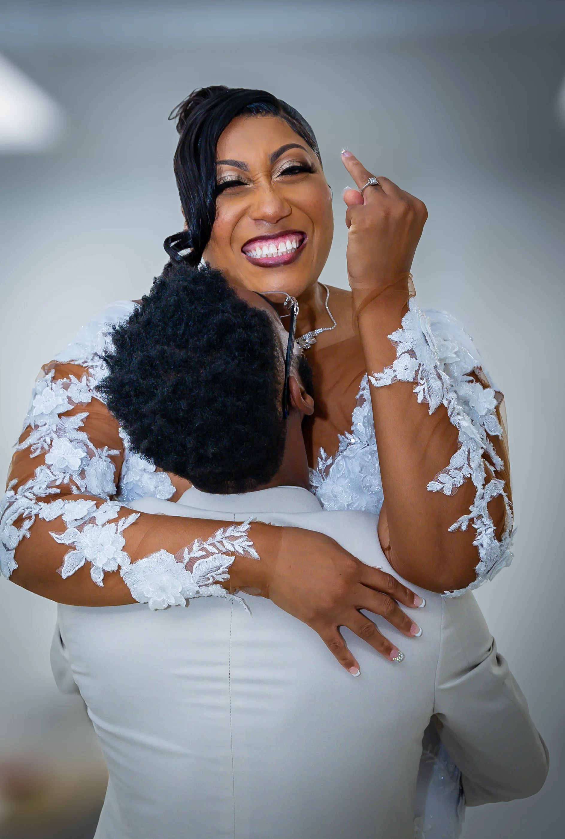 A woman in a white lace dress smiling and proudly pointing her finger, being embraced by a person whose face is not visible.