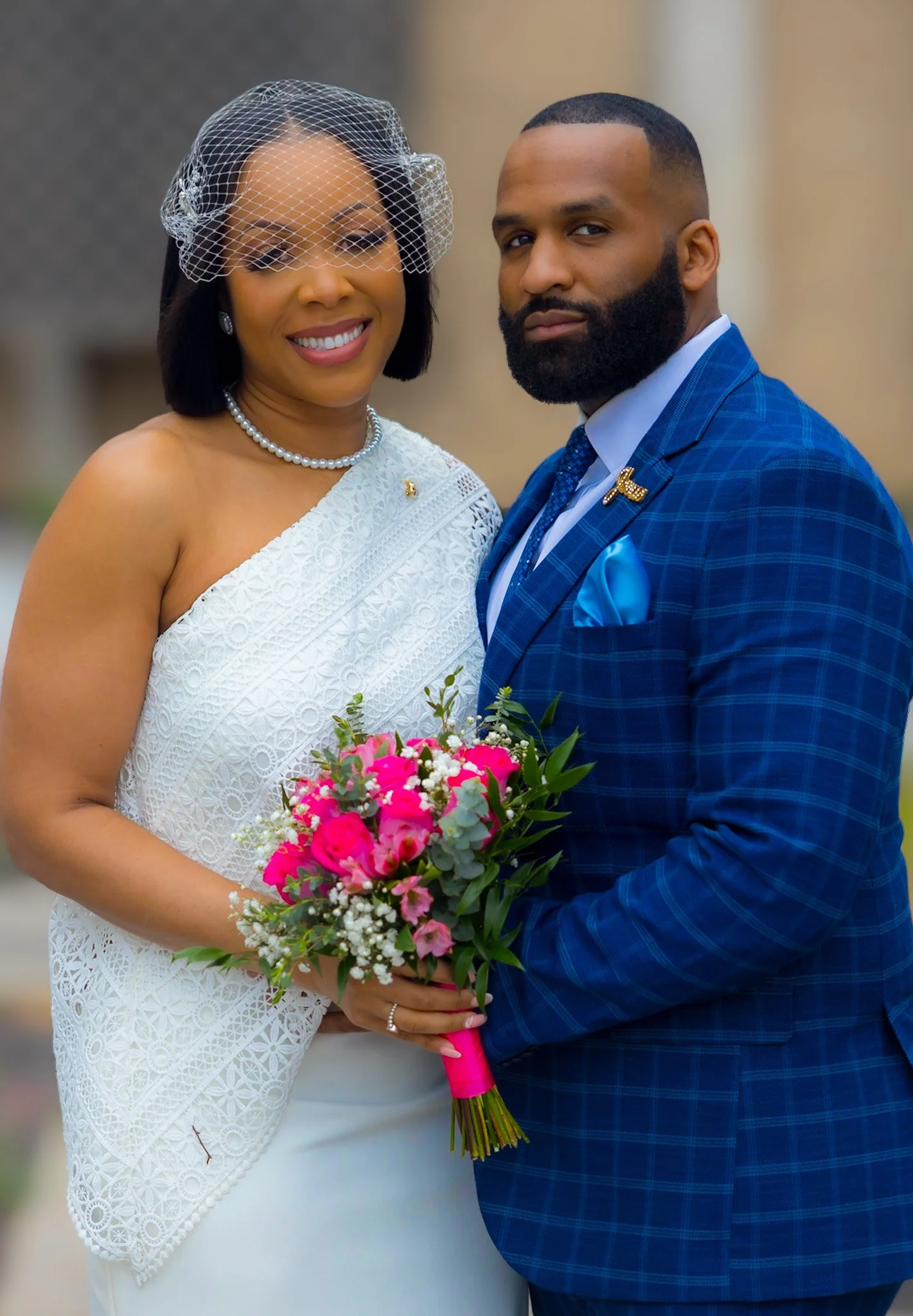 A woman in a white dress with a lace pattern holding a bouquet of pink flowers, standing beside a man in a blue checkered suit with a blue pocket square and a pin, outdoor background.