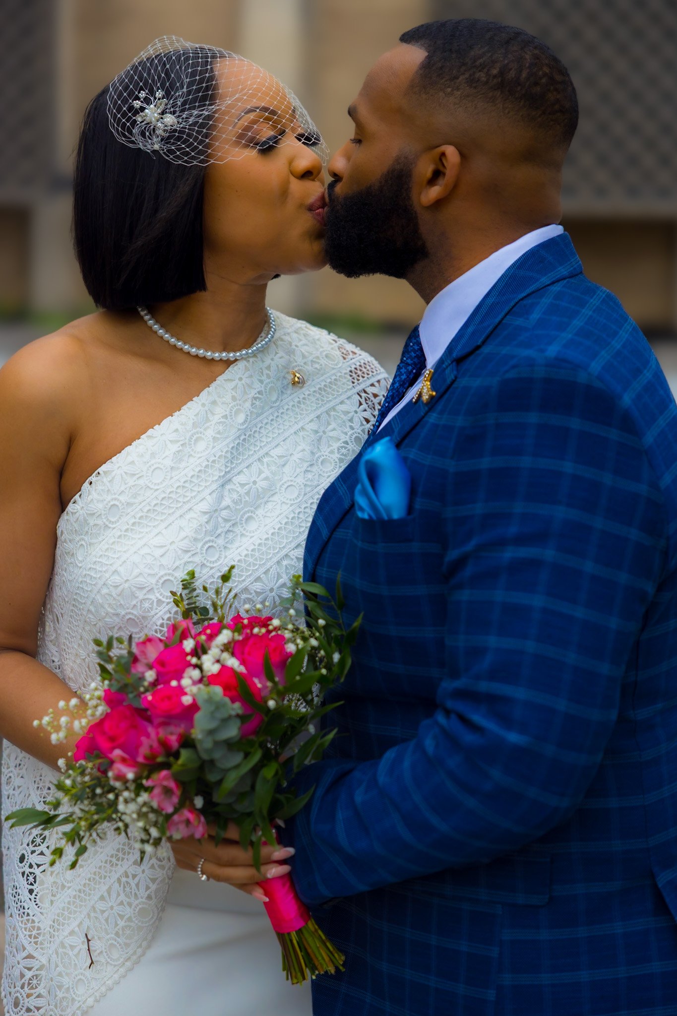 A couple on their wedding day, sharing a kiss. The woman wears a white lace dress and pearl necklace, holding a bouquet of pink roses and greenery. The man wears a blue checkered suit with a blue pocket square.