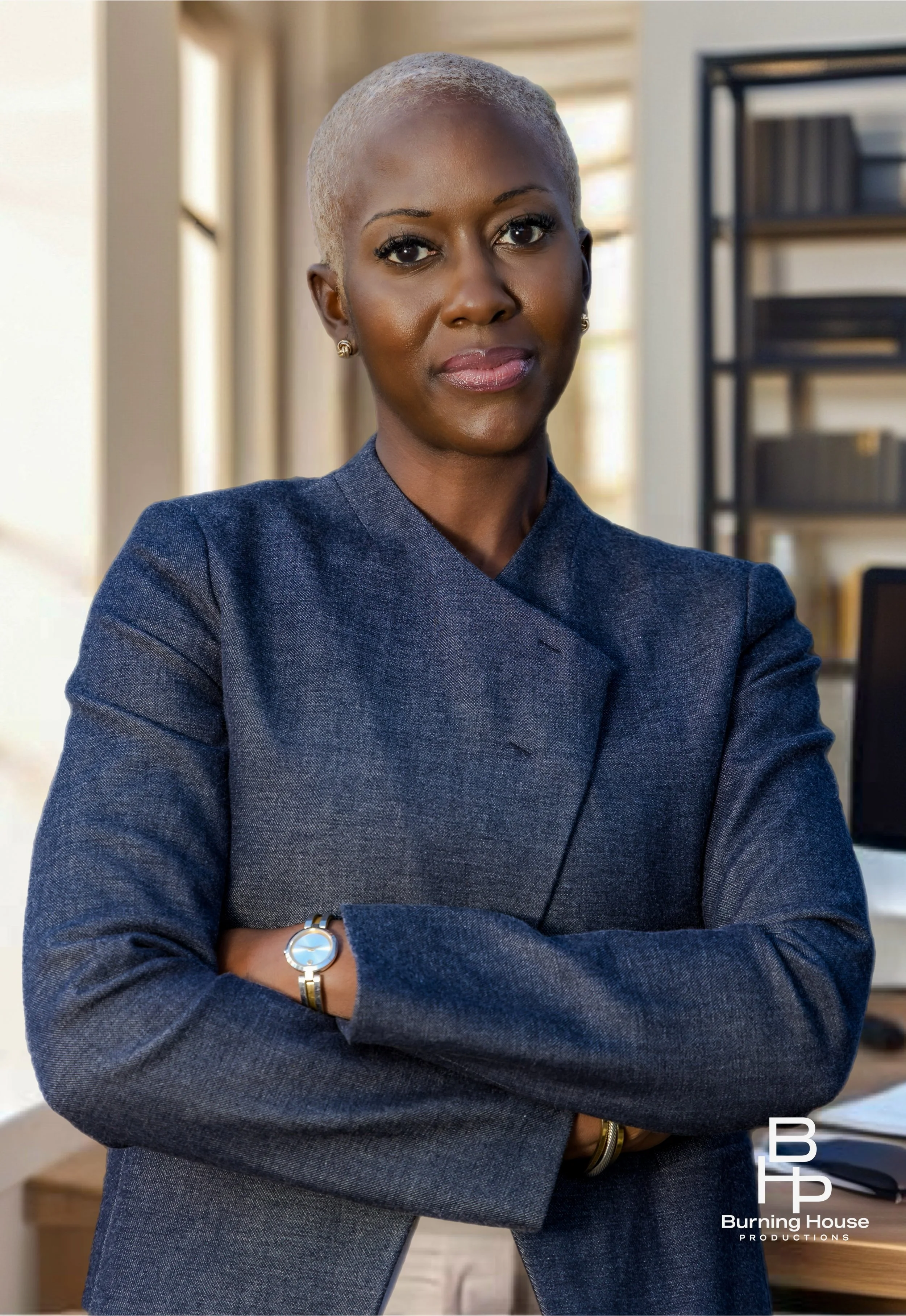 A confident woman with short platinum blonde hair in a navy blazer standing with arms crossed in an office setting.
