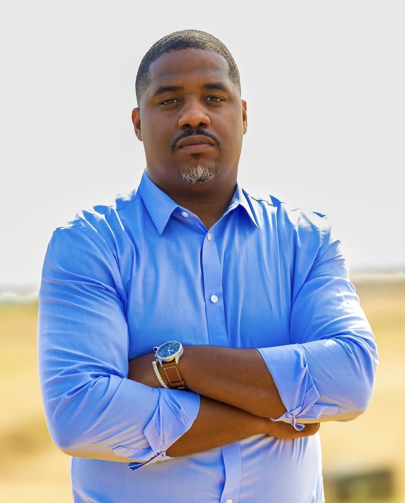 A man standing outdoors with arms crossed wearing a light blue button-up shirt and a watch, looking directly at the camera.
