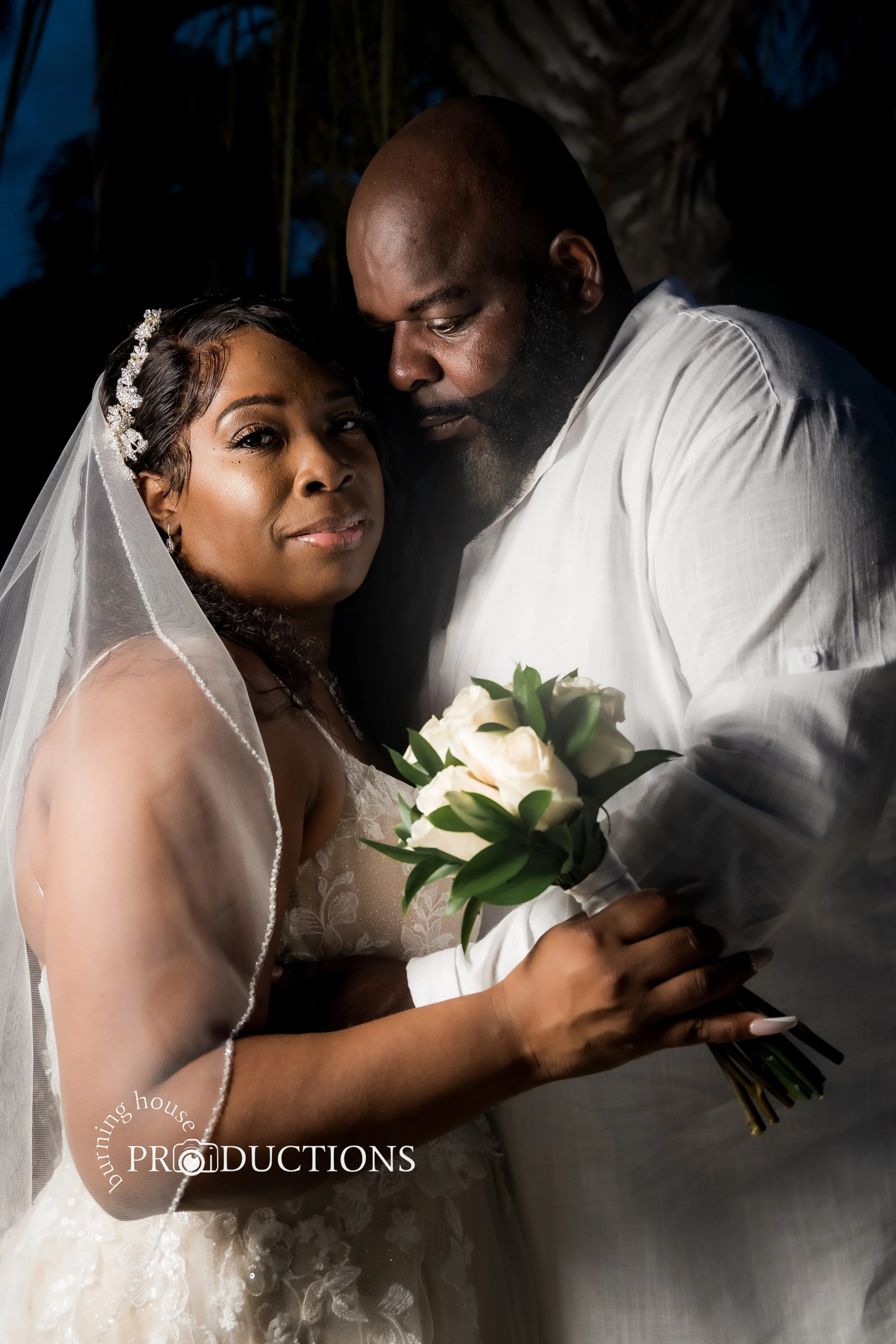 A bride and groom pose closely together during their wedding. The bride is holding a bouquet of white roses, and they are both dressed in white wedding attire. The bride has focus on the camera, while the groom's face is angled down towards her.