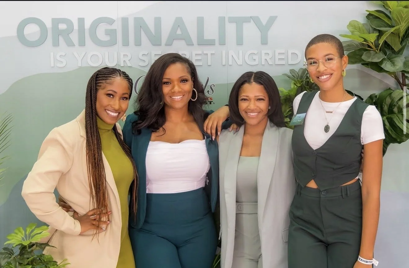 Four women standing together in front of a sign that reads "Originality is your secret ingredient." They are smiling, dressed in business casual attire, with plants in the background.
