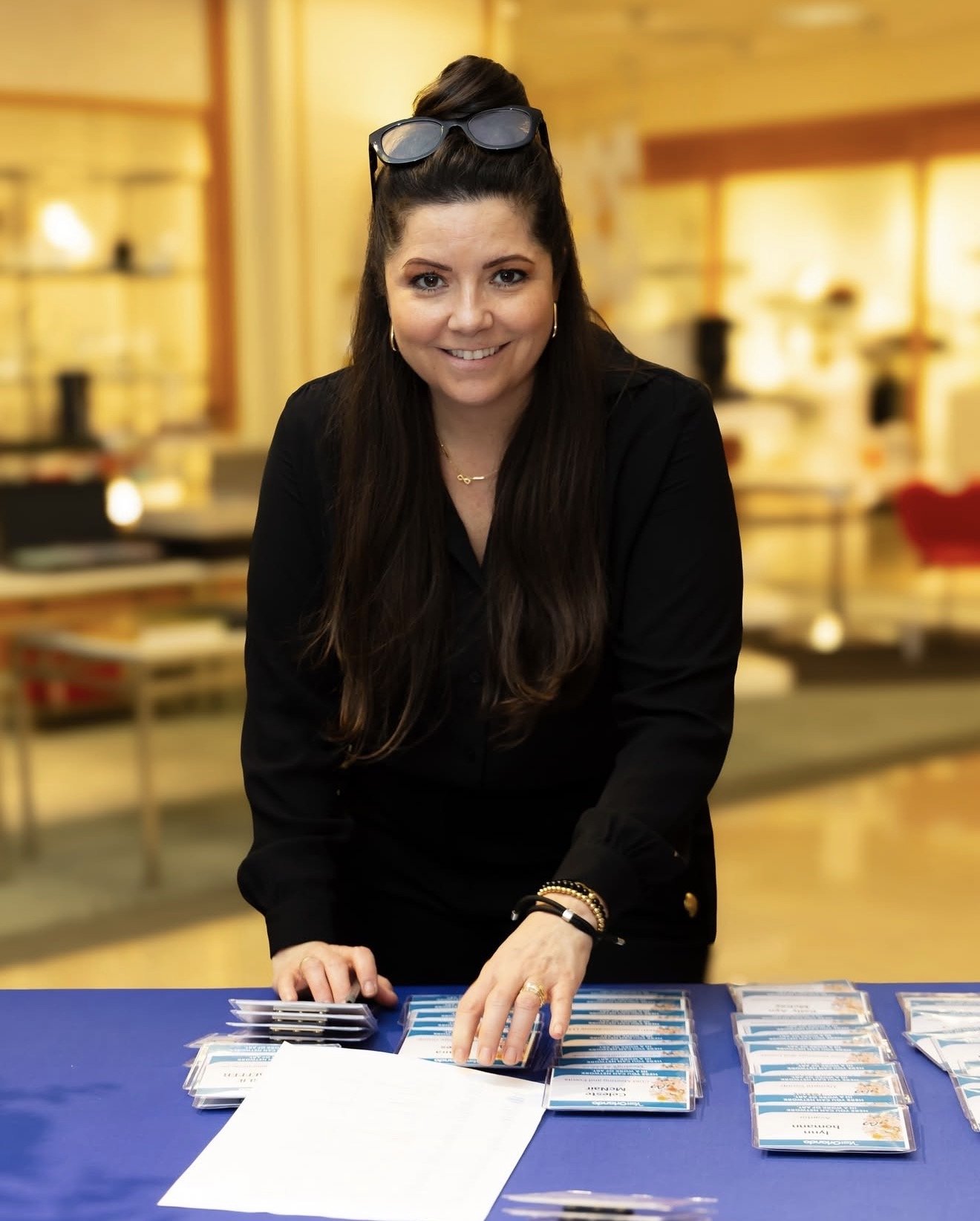 A woman with long dark hair, wearing a black outfit and sunglasses on her head, is smiling and organizing name badges or event passes on a table with a blue tablecloth.