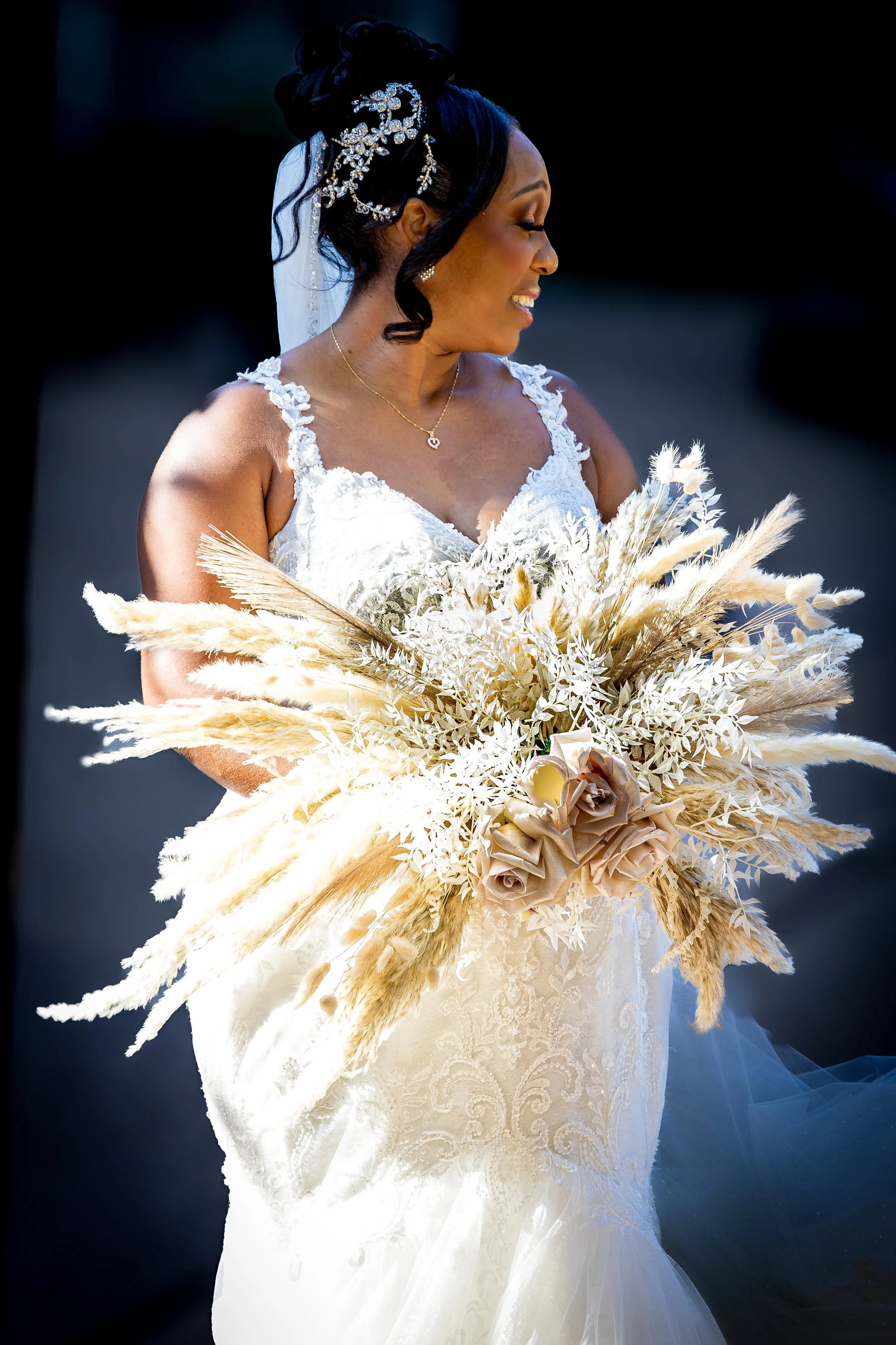A smiling bride in a white wedding dress holding a large bouquet of dried flowers and wheat, with dark hair styled in an updo decorated with a flower and pearl hair accessory.