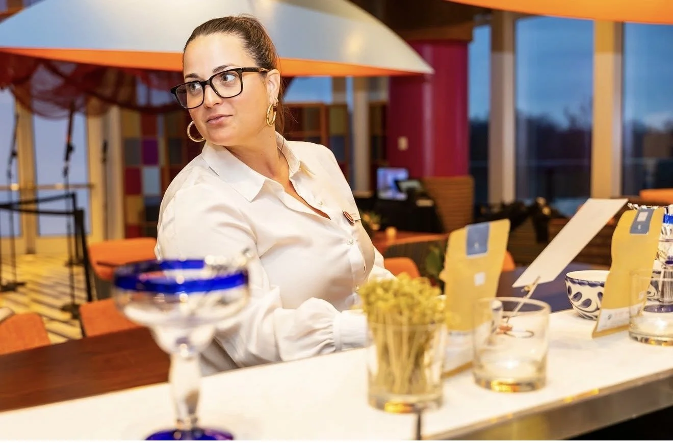 A woman with dark hair tied in a bun, wearing glasses, earrings, and a white shirt, sitting at a counter in a colorful restaurant or cafe with tables, chairs, and large windows.