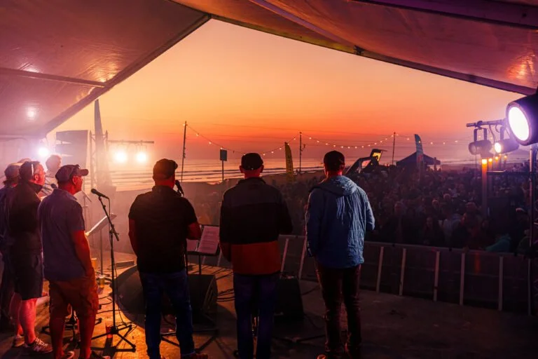 Group of musicians performing on stage at an outdoor concert during sunset, with a large audience in front and colorful tents overhead.