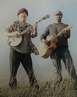 Two men standing outdoors in a grassy field, playing guitars and banjos.