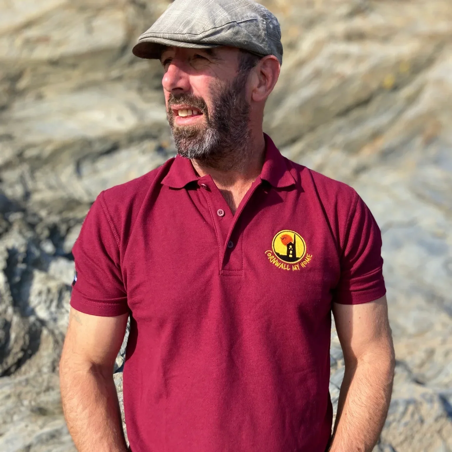 A man with a beard wearing a gray flat cap and a maroon polo shirt with a 'Cornwall My Home' logo, standing outdoors in front of a rocky background.