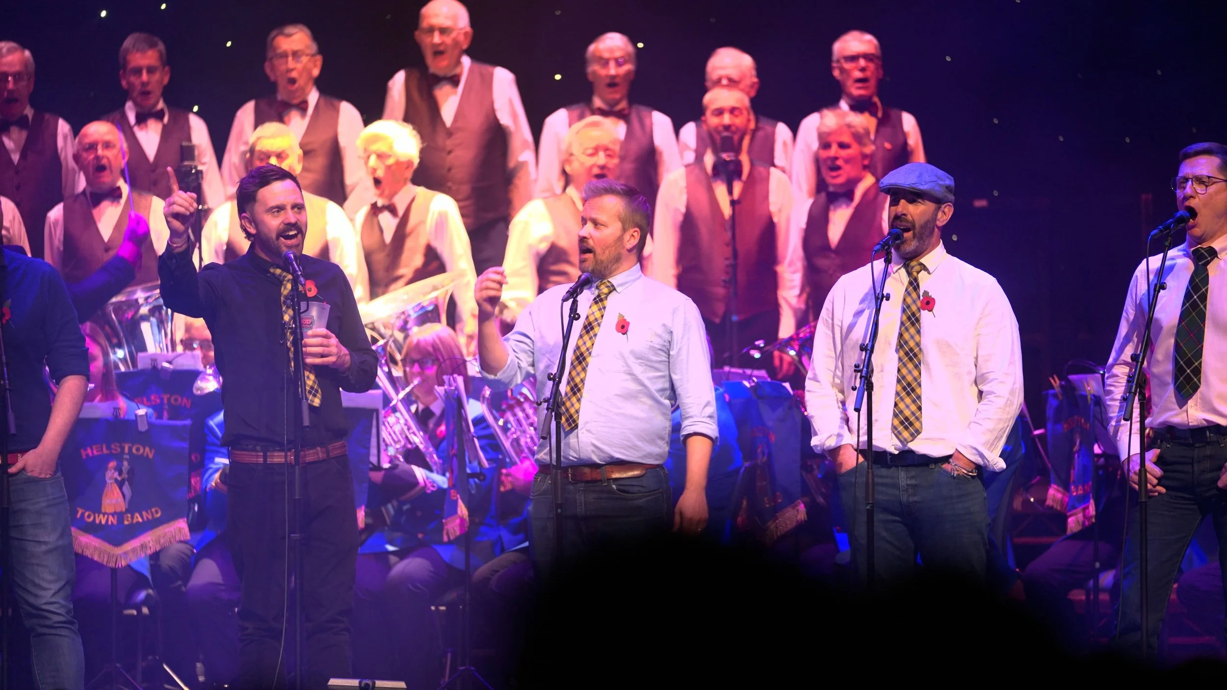 A group of men and women performing on stage, with some singing into microphones and others sitting or standing in the background, wearing formal and semi-formal attire including ties and vests, in a concert setting with musical instruments and banne